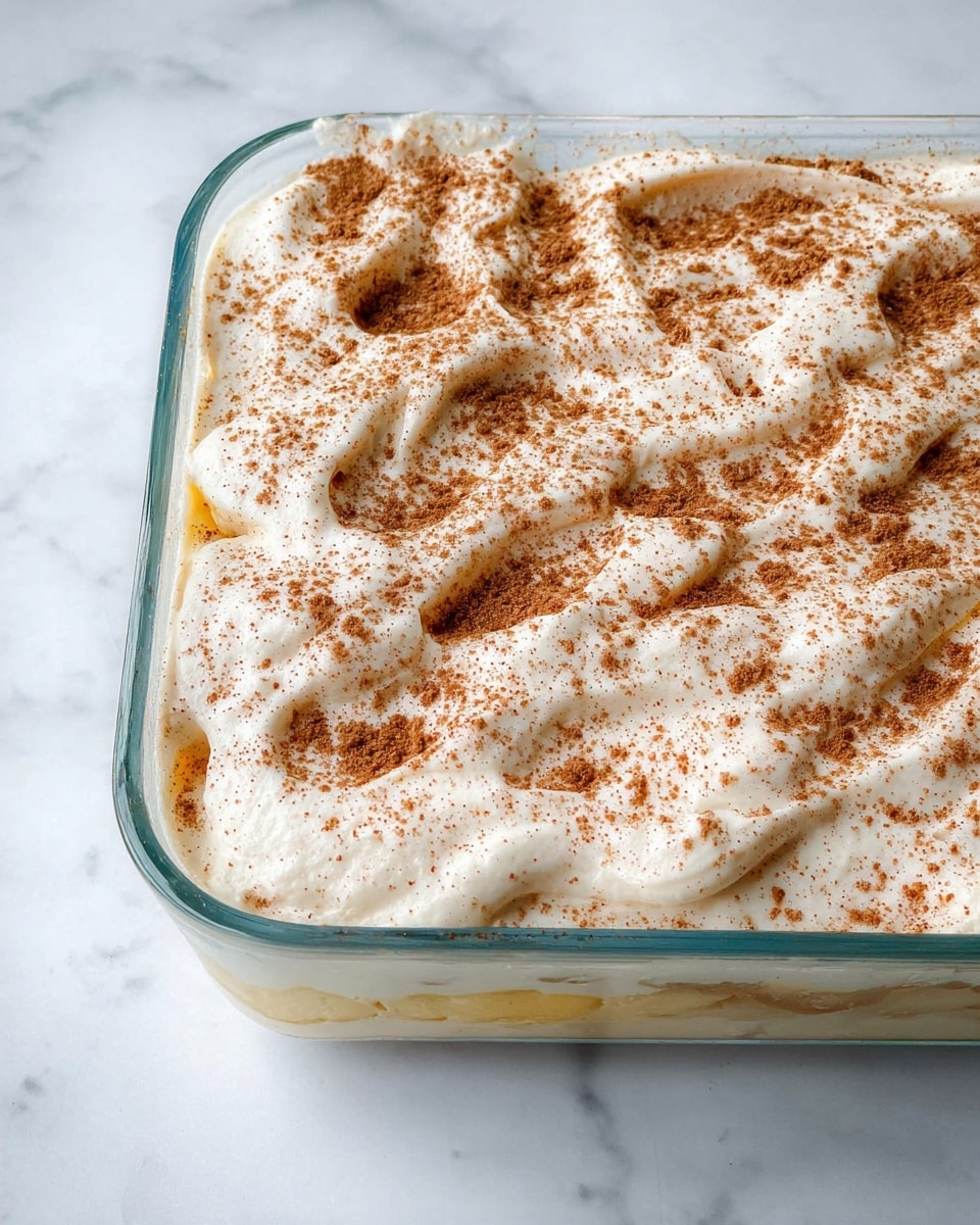 A glass rectangular baking dish contains a dessert made up of two visible layers: a thick, creamy white top layer with a slightly uneven, whipped texture sprinkled evenly with light brown cinnamon powder, sitting over a thin, pale yellow base layer beneath it. The dish is placed on a white marbled surface. Photo taken with an iphone --ar 4:5 --v 7