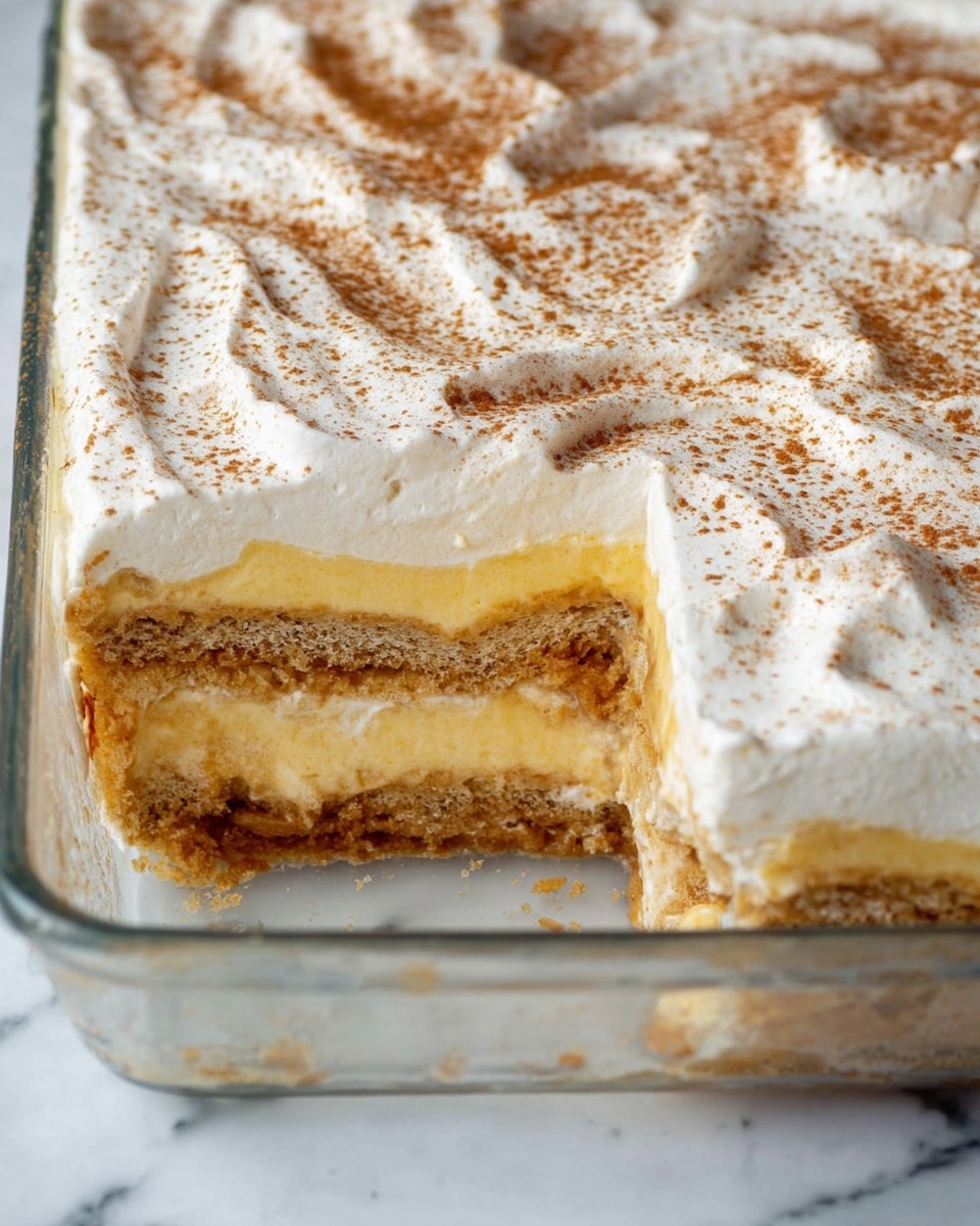 The image shows a multi-layered dessert in a clear rectangular glass dish against a white marbled surface. The dessert has four visible layers: the bottom layer appears to be a soaked, light brown sponge or biscuit base, followed by a thick pale yellow creamy layer, topped by another soaked sponge layer, and then a final thick yellow cream layer. The top of the dessert is covered with a smooth white whipped cream layer, which has some soft swirls and is lightly sprinkled with a fine brown powder, likely cinnamon or cocoa. A large section is missing from the front left of the dish, revealing the layered inside clearly. Photo taken with an iphone --ar 4:5 --v 7