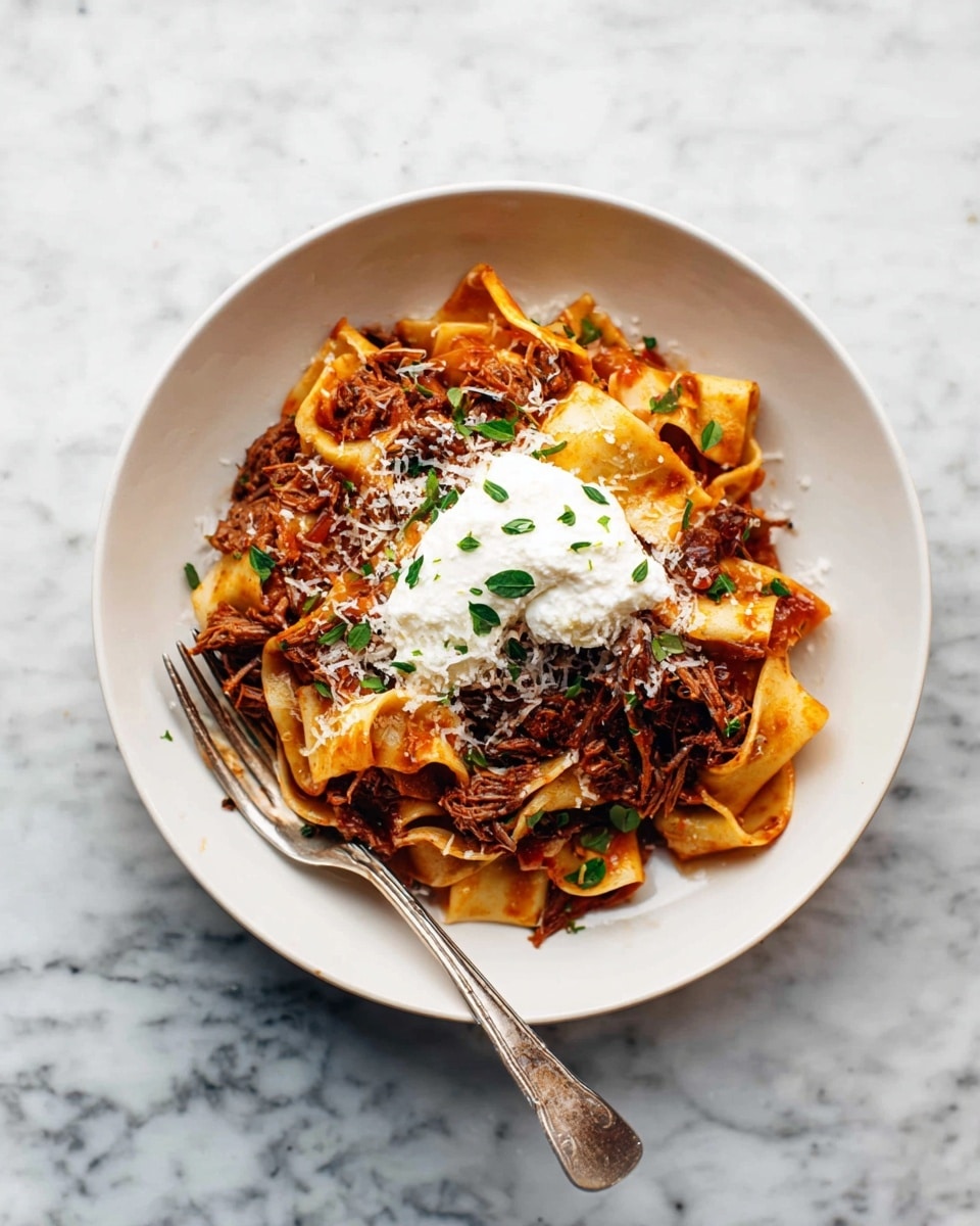A white plate holds wide, flat yellow pasta ribbons mixed with shredded brown meat in a rich red sauce. The pasta and meat are topped with crumbled white cheese and green herb leaves, with a dollop of white creamy cheese placed in the center. A silver fork is resting on the left side of the plate. The plate sits on a white marbled surface. photo taken with an iphone --ar 4:5 --v 7