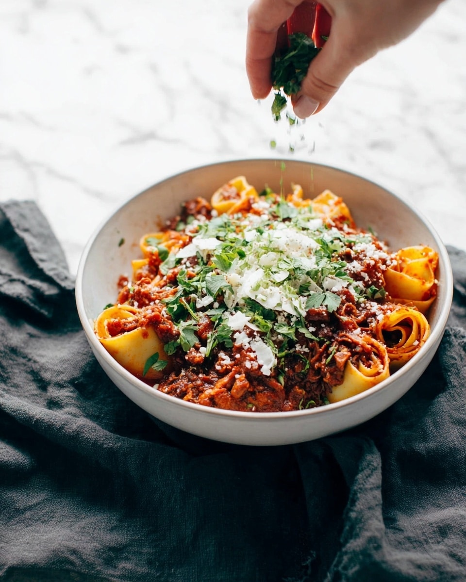 A white bowl is filled with a layered pasta dish, starting with a base of rolled pasta tubes stuffed with shredded meat. The pasta is covered with a rich red sauce that has visible bits of meat and vegetables. On top, there is a sprinkle of white cheese and fresh green herbs. A woman's hand is sprinkling more fresh green herbs over the dish from above. The bowl sits on a dark cloth, but the background is a white marbled texture. photo taken with an iphone --ar 4:5 --v 7