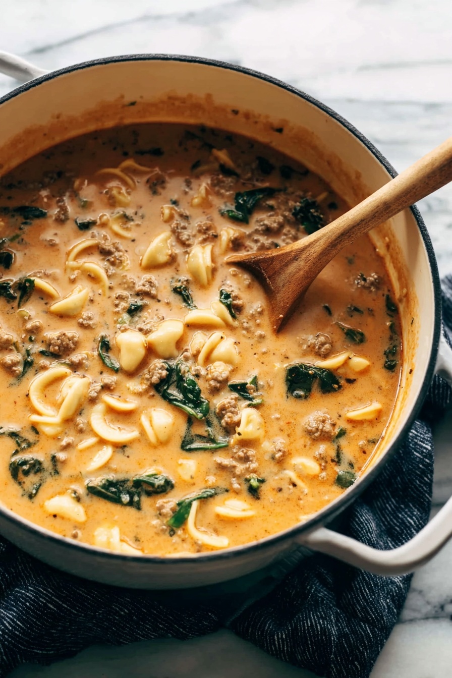 Creamy Tortellini Soup with Italian Sausage, Italian sausage tortellini soup, hearty creamy tortellini soup, easy Italian sausage pasta soup, comforting tortellini soup recipes - A large white pot filled with a thick, creamy soup that is orange-beige in color, speckled with small pieces of ground meat, green leafy vegetables, and small curved pasta shells. A wooden spoon is resting inside the pot, partially submerged in the soup, showing its rich and smooth texture. The pot sits on a dark blue cloth on a white marbled surface, creating a warm and cozy feeling. photo taken with an iphone --ar 2:3 --v 7