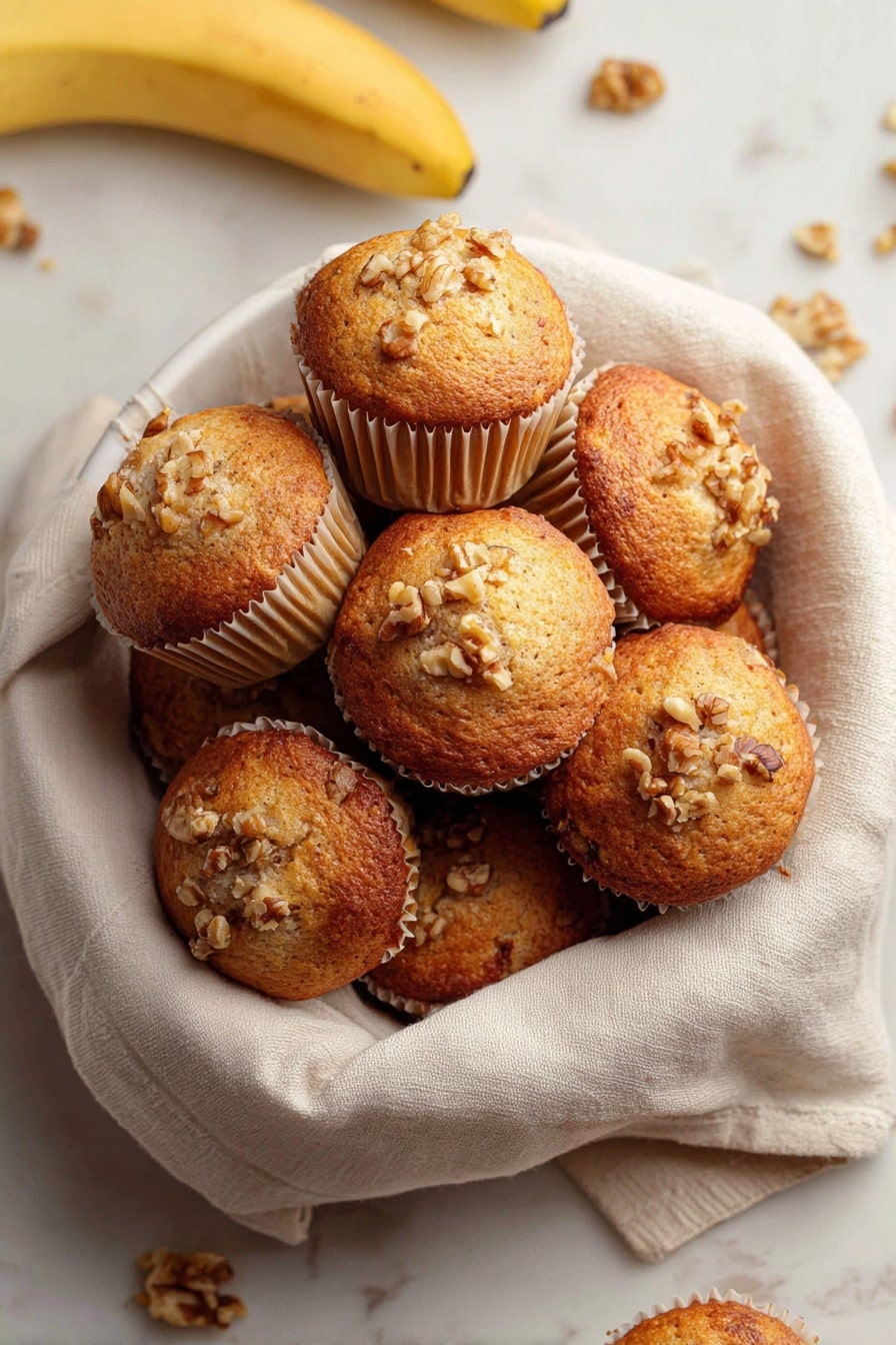Quick & Easy Vegan Banana Muffins, vegan banana muffins, healthy banana muffin recipe, gluten-free banana muffins, breakfast banana muffins - A white bowl lined with a soft beige cloth holds a pile of golden brown muffins with slightly domed tops. Each muffin is in light beige paper cups and sprinkled with small pieces of walnuts on the top, giving a rough texture. Around the bowl on a white marbled surface, there are a few loose walnuts and bananas visible, adding natural color contrasts. The muffins appear moist with a lightly browned crust. photo taken with an iphone --ar 2:3 --v 7