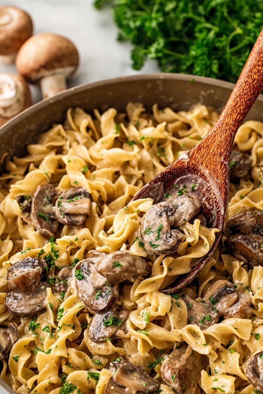 Old Fashioned Beef Stroganoff, Beef Stroganoff recipe, classic beef stroganoff, homemade beef stroganoff, easy beef stroganoff - The image shows a close-up of a pan filled with a creamy noodle and mushroom dish. There are egg noodles that are light yellow, soft, and curly, spread all over the pan as the first layer. On top of the noodles, there are sliced mushrooms with a brown color and slightly shiny surface from the sauce. Mixed with the mushrooms and noodles, there are small bits of green parsley scattered for color. A wooden spoon is scooping some of the dish from the pan. The background has some blurred green parsley and whole mushrooms placed on a white marbled surface. photo taken with an iphone --ar 2:3 --v 7