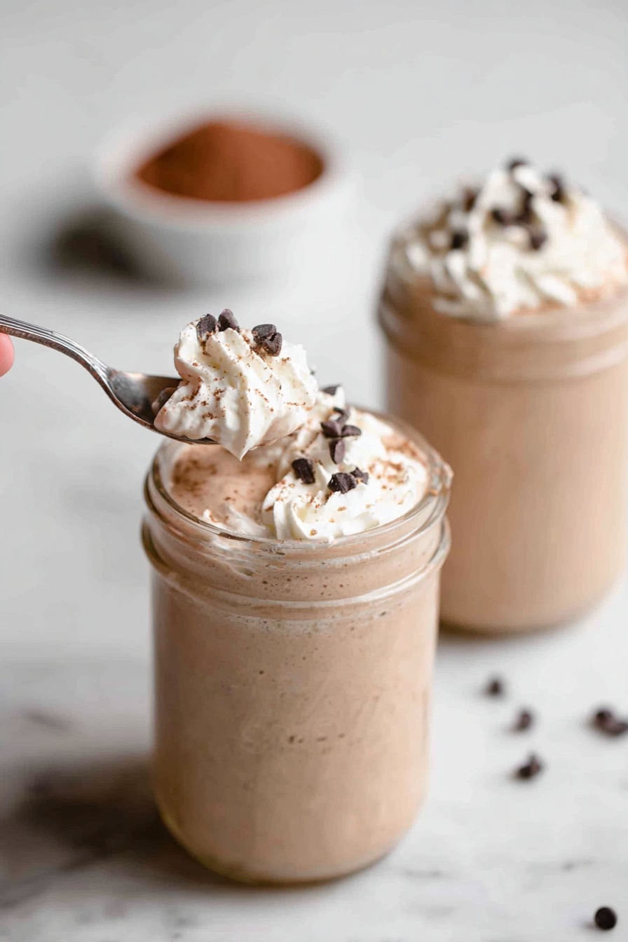 Vegan Chocolate Banana Protein Shake, healthy vegan smoothie, quick plant-based protein shake, chocolate banana smoothie, nutritious vegan breakfast - Two glass jars sit on a white marbled surface, each filled with a creamy light brown milkshake. The milkshakes have a thick texture and are topped with a swirl of white whipped cream. Small, dark chocolate chips are sprinkled over the whipped cream. In the foreground, a silver spoon held by a woman's hand scoops a portion of whipped cream and chocolate chips from the first jar. The background jar is out of focus but shows the same whipped cream and chocolate chip topping. There is a small bowl filled with cocoa powder blurred in the background. Photo taken with an iphone --ar 2:3 --v 7