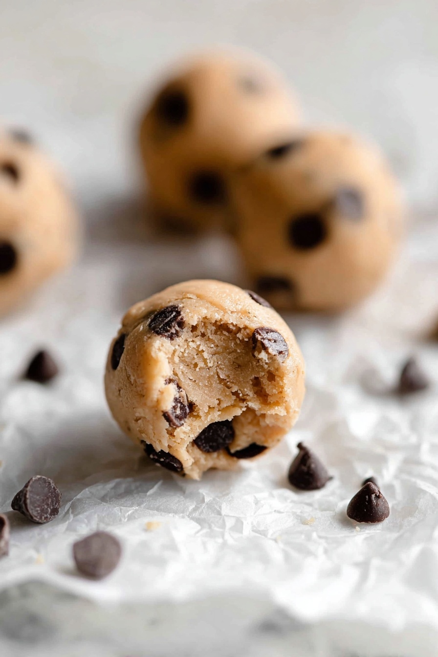 Protein Cookie Dough Bites, healthy protein snacks, no-bake protein treats, high-protein dessert ideas, easy healthy snack recipes - The image shows small round cookie dough balls with a light tan color, filled with many dark brown chocolate chips scattered inside and on the surface. The balls rest on a crumpled white paper on a white marbled background. One dough ball in the front has a small bite taken out of it, showing a soft, slightly crumbly texture inside, while other dough balls and some loose chocolate chips are blurred in the background. The lighting is soft and natural, highlighting the texture of the dough and chocolate chips, photo taken with an iphone --ar 2:3 --v 7