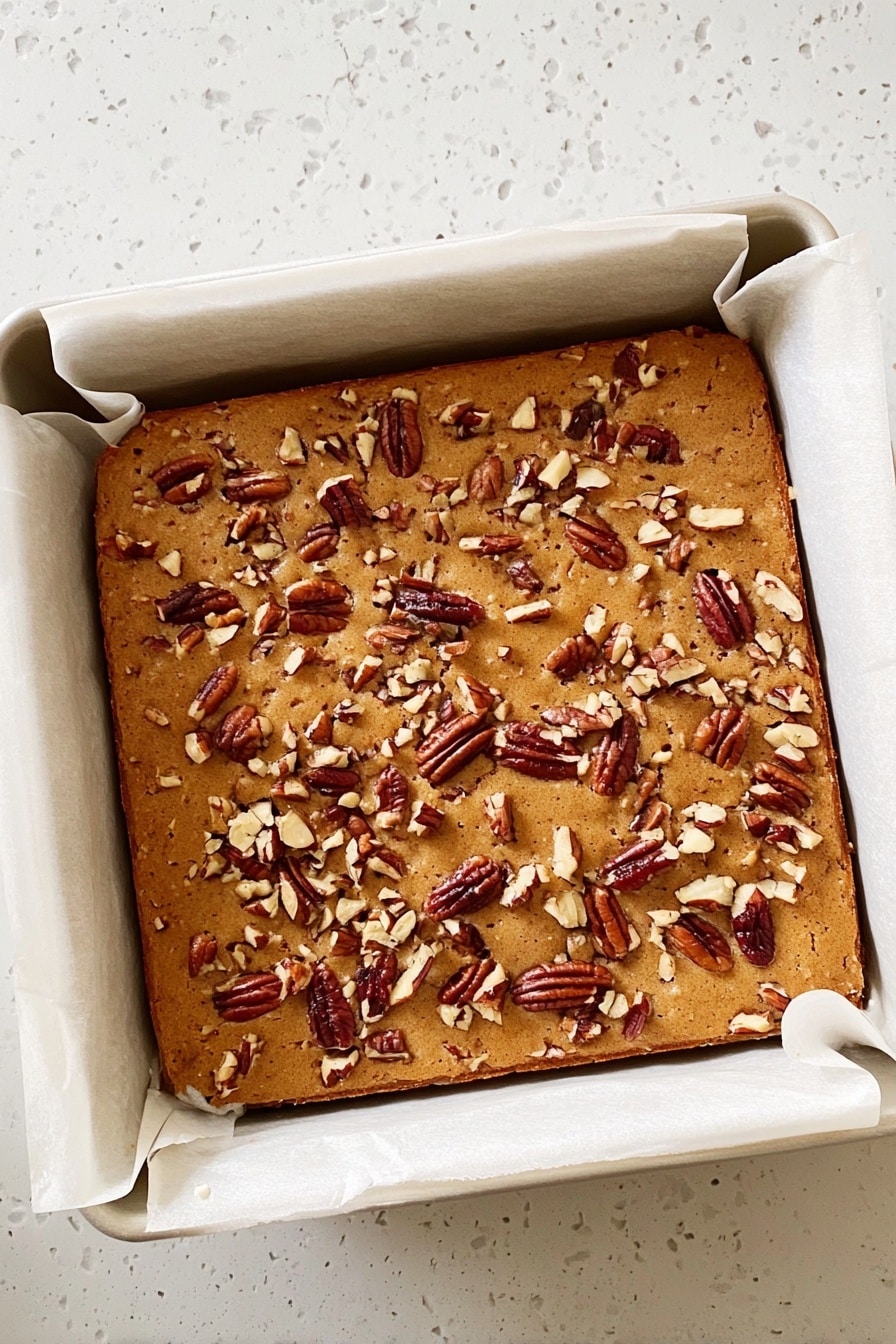 Maple Pecan Blondies, Maple Pecan Blondies recipe, how to make blondies, easy blondie desserts, nutty maple blondies - A square baked dessert with a light brown top covered in scattered pieces of pecans. It is in a white baking pan lined with parchment paper that folds over the edges. The baked item looks soft and slightly puffy, with a smooth but slightly textured surface from the nuts. The pan sits on a white marbled surface. photo taken with an iphone --ar 2:3 --v 7