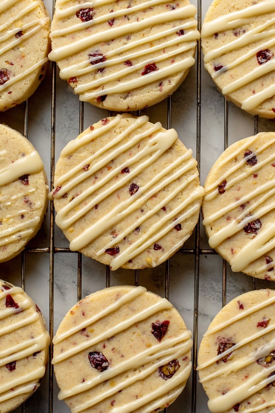 Orange Cranberry Shortbread Cookies, citrus cranberry cookie recipe, easy holiday shortbread, buttery fruit cookies, cranberry orange dessert - A close-up view of a batch of round cookies laid out on a metal cooling rack over a white marbled surface. Each cookie has one visible layer, light golden in color with small dark red and black fruit bits inside, giving a spotted look. On top of each cookie is a thin, even drizzle of pale cream-colored icing, applied unevenly in long lines across the surface. The texture of the cookies looks soft but firm, with some slight cracks near the edges. The metal wires of the rack create a grid pattern underneath the cookies. Photo taken with an iphone --ar 2:3 --v 7