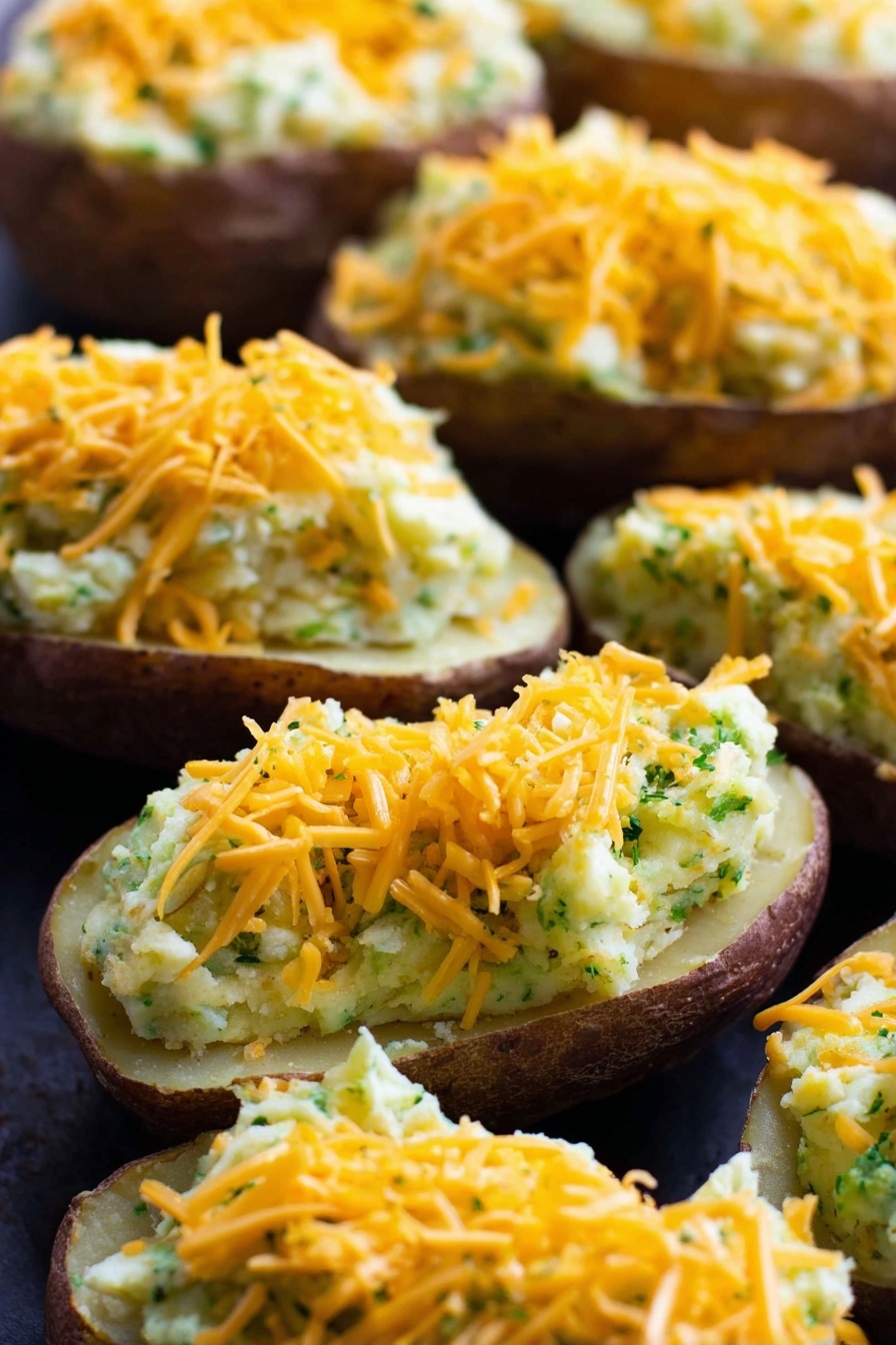 Vegan Cheesy Broccoli Twice Baked Potatoes, vegan potato recipes, plant-based stuffed potatoes, dairy-free cheesy potatoes, healthy vegan potato dishes - The image shows several potato halves with the inside scooped out and filled with a creamy mixture that looks light yellow with green bits throughout, suggesting herbs or vegetables mixed in. Each stuffed potato is topped with a generous layer of shredded orange cheddar cheese. The potatoes are arranged closely together on a dark surface which contrasts with the light filling and bright cheese. The texture of the filling appears smooth but slightly chunky, and the potato skins are a dark brown color. The close-up shot highlights the layers clearly with the cheese on top, the creamy filling in the middle, and the potato skin at the bottom. Photo taken with an iphone --ar 2:3 --v 7