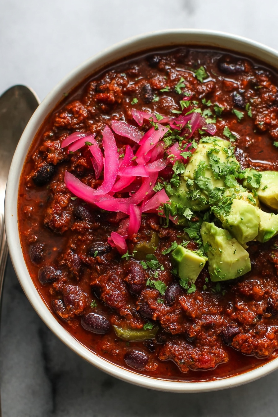 Lentil Chili, healthy lentil chili, vegetarian chili, vegan chili recipe, easy lentil chili - A close-up view of a bowl of thick chili, showing three main layers: a base layer of rich, dark red-brown chili with black and red beans and small pieces of green bell pepper spread throughout, topped on one side with bright pink pickled onion slices, and on the same side, fresh green avocado chunks sprinkled with finely chopped cilantro. The bowl is white and a spoon is partially visible on the left side, all placed on a white marbled surface. Photo taken with an iphone --ar 2:3 --v 7