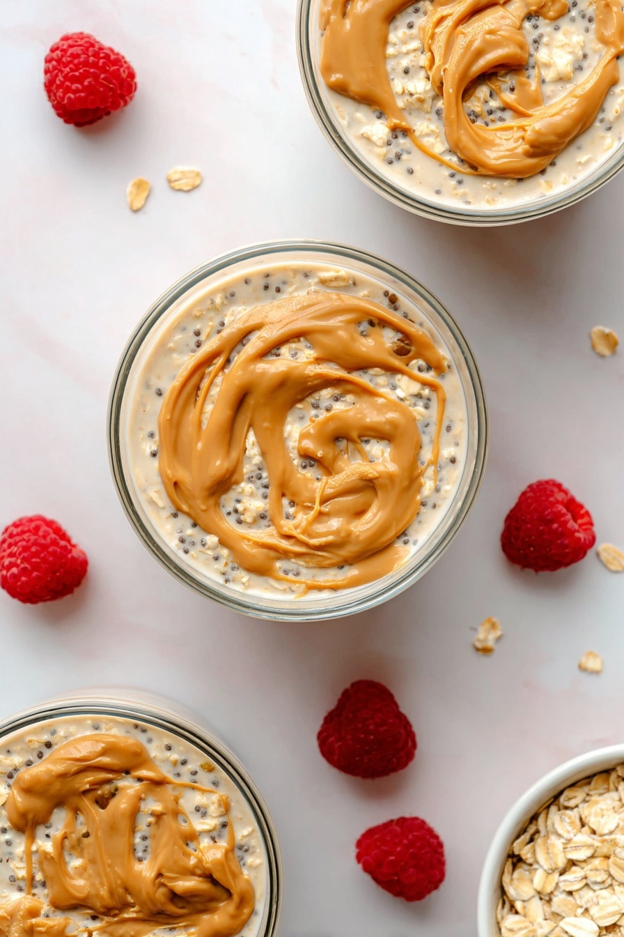 Peanut Butter Overnight Oats, healthy overnight oats, easy breakfast recipes, quick morning breakfast, nutritious oatmeal - The image shows three round clear glass bowls filled with layered overnight oats on a white marbled surface. Each bowl has a bottom layer of creamy light beige oats mixed with small black chia seeds, covered by a thin layer of milky liquid. The top layer is swirled with dollops of light brown peanut butter, creating smooth, curved textures on the surface. Around the bowls, scattered fresh bright red raspberries add a pop of color, along with a small white bowl filled with rolled oats placed in the bottom right corner. The photo taken with an iphone --ar 2:3 --v 7