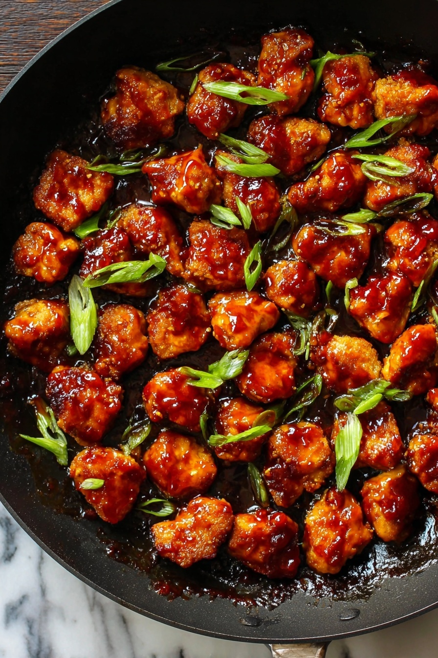 Honey Ginger Seitan with Crispy Pan-Fried Coating, plant-based seitan recipes, vegan crispy seitan, Japanese-inspired vegan dishes, healthy plant protein dishes - This image shows a black pan filled with many small pieces of golden-brown chicken covered in thick, shiny reddish-brown sauce. The pieces are spread out evenly across the pan, with some bright green slices of scallion sprinkled on top. The sauce looks sticky and glossy, covering all the chicken pieces in a rich layer. The pan's dark color contrasts with the vibrant colors of the chicken and scallions. The background surface is a white marbled texture. photo taken with an iphone --ar 2:3 --v 7