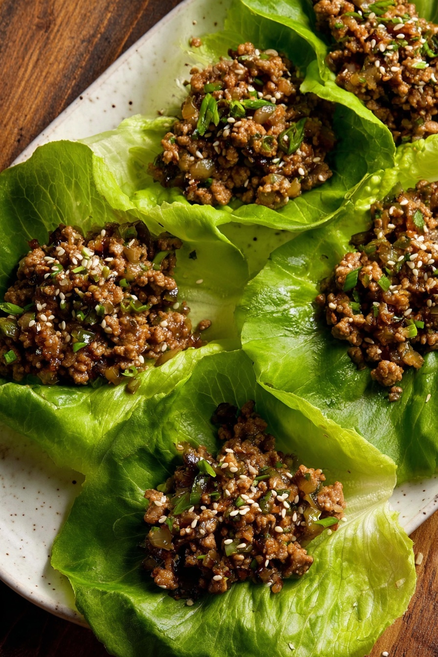 Tofu Lettuce Wraps with Mushrooms and Water Chestnuts, healthy tofu lettuce wraps, vegetarian lettuce wraps with mushrooms, crunchy water chestnuts appetizer, easy tofu lettuce wraps - The image shows five large green lettuce leaves arranged on a white plate with a wood surface in the background. Each leaf holds a serving of cooked ground meat mixed with small diced vegetables, with a brown color and a slightly moist texture. The meat mixture is sprinkled with white sesame seeds and small bits of green herbs. The lettuce leaves are fresh with visible veins and crinkled edges, creating a bright green contrast to the darker meat filling. Photo taken with an iphone --ar 2:3 --v 7