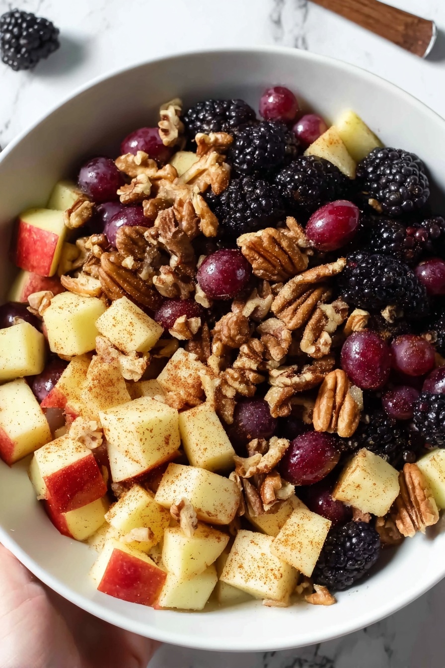 Fall Fruit Salad with Maple Cinnamon Dressing, autumn fruit salad, healthy fall fruit salad recipe, cinnamon maple salad, seasonal fruit salad - A white bowl is filled with a fruit salad that has three main layers: the bottom layer is made of blackberries, which are bumpy and dark purple-black; the middle layer has round red grapes with smooth, shiny skin; and the top layer contains apple chunks that are pale yellow with a dusting of brown cinnamon powder. Scattered throughout the salad are light brown pecans adding a rough texture. The bowl sits on a white marbled surface, and two cinnamon sticks lie nearby. photo taken with an iphone --ar 2:3 --v 7