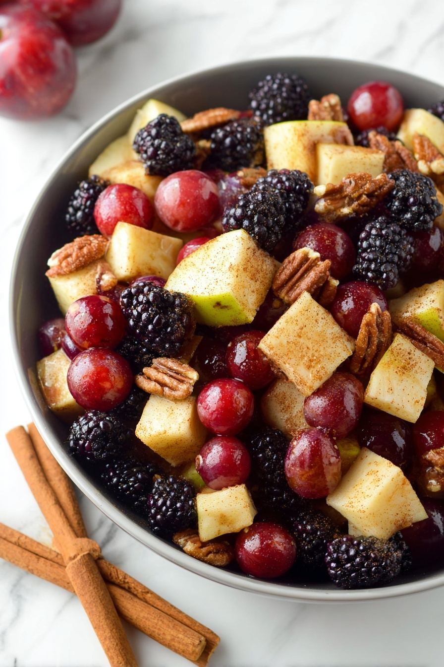 Fall Fruit Salad with Maple Cinnamon Dressing, autumn fruit salad, healthy fall fruit salad recipe, cinnamon maple salad, seasonal fruit salad - A white bowl filled with a colorful fruit and nut salad, showing three layers of ingredients. The bottom layer is pieces of pale yellow apple with red skin, cut into small cubes. The middle layer features deep red grapes and dark purple-black blackberries scattered evenly across. The top layer includes crunchy brown walnuts and pecans mixed throughout. The fruit pieces have a light dusting of cinnamon or spice, giving a slight matte texture. The bowl is placed on a white marbled surface, and a woman's hand is visible holding the bowl's edge. photo taken with an iphone --ar 2:3 --v 7