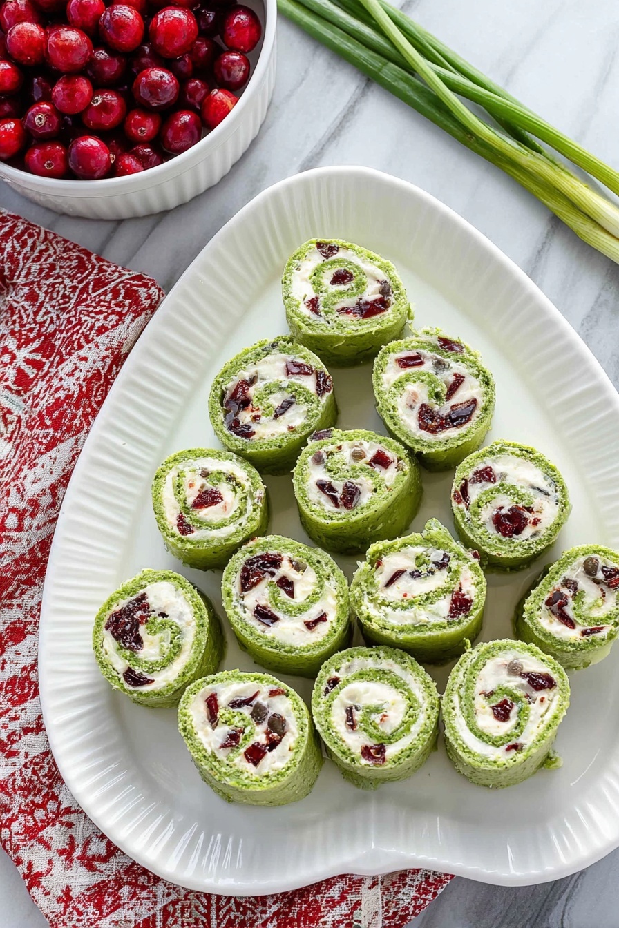 Cranberry Feta Pinwheels, Cranberry Feta Wraps, Easy Cranberry Appetizer, Holiday Cranberry Snacks, Cream Cheese Pinwheels - On a white oval plate with a ridged edge, there are fifteen green pinwheel rolls arranged in a triangular shape, with one at the top, three in the second row, five in the third row, and six in the bottom row. Each roll shows a swirl of bright green outer layer with a creamy white filling inside that contains small pieces of dark red and green ingredients mixed throughout. The plate is placed on a white marbled surface, and to the top left, a white bowl filled with red berries is partially visible, sitting on a red and white patterned cloth. A few green onion stems lie on the top right corner of the surface. Photo taken with an iphone --ar 2:3 --v 7