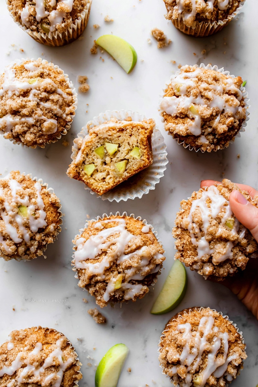 Apple Cinnamon Crumb Muffins, apple cinnamon muffins recipe, cinnamon apple muffins, crumb muffin recipe, cozy breakfast muffins - The image shows nine apple crumb muffins arranged on a white marbled surface. Each muffin has three clear layers: a bottom baked golden-brown cake layer, a middle layer with visible light green apple chunks inside the muffin, and a top crumbly light brown streusel topping with a slight sprinkle of white glaze drizzled unevenly over it. One muffin is open, showing the inside texture and apple pieces, sitting in a white paper liner with a woman's hand about to pick it up. Small crumbs and a few green apple slices are scattered around, adding detail to the scene. Photo taken with an iphone --ar 2:3 --v 7