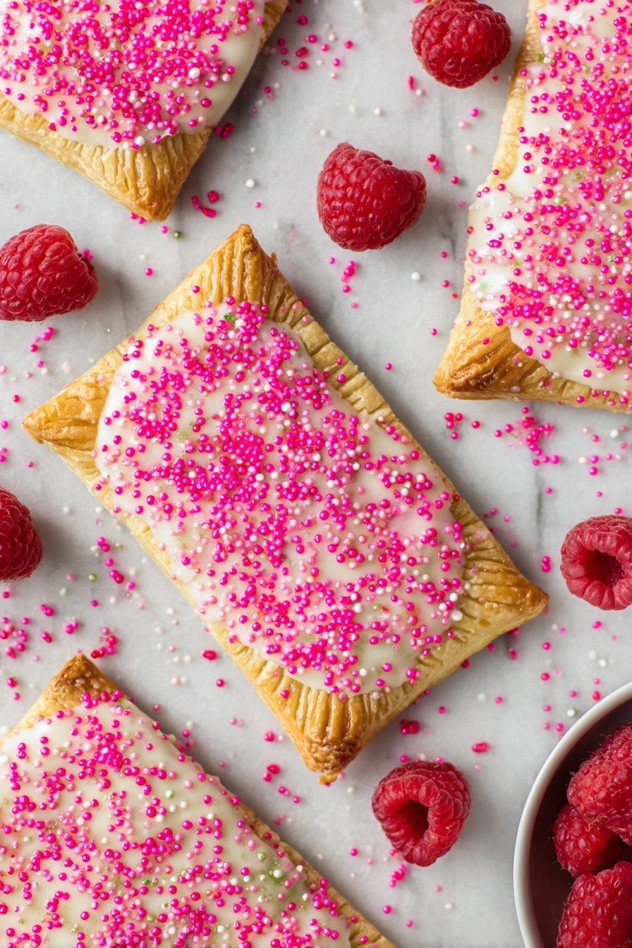 Raspberry Pop Tarts with Vanilla Icing, homemade raspberry pop tarts, flaky fruit-filled breakfast pastry, easy homemade pop tarts, vanilla icing for pastries - Several rectangular toasted pastries spread over a white marbled surface, each with a golden-brown crust stamped with fork marks along the edges; the top layer is coated in a slightly shiny white icing sprinkled generously with bright pink sugar crystals, creating a textured look. Scattered around the pastries are fresh, plump red raspberries adding a pop of natural color. In the bottom right corner, a small portion of a white bowl is visible, holding more raspberries. photo taken with an iphone --ar 2:3 --v 7