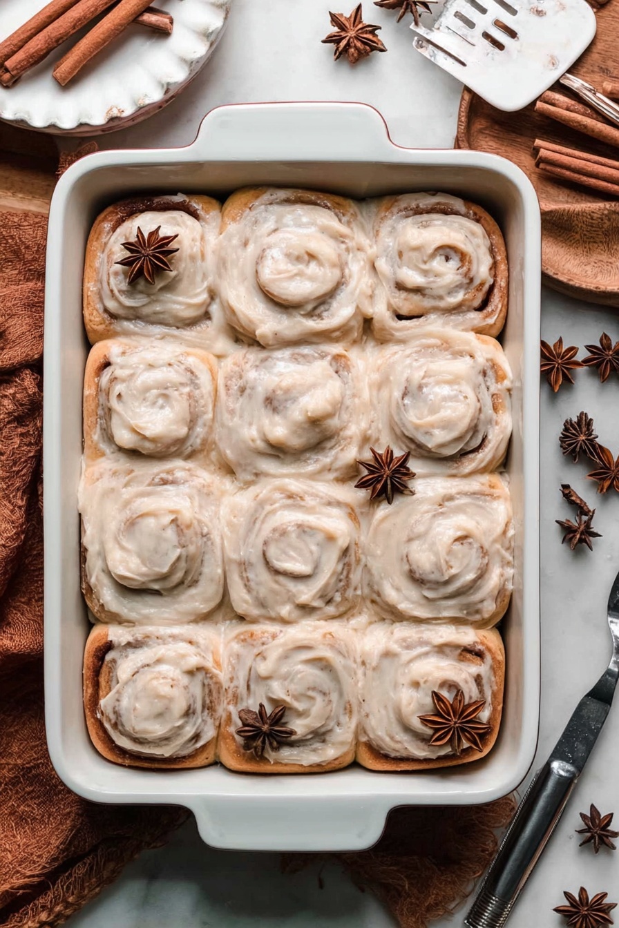 Chai Cinnamon Rolls with Caramel Drizzle, chai cinnamon roll recipe, spiced cinnamon rolls, caramel cinnamon roll dessert, cozy chai breakfast - A white rectangular baking dish filled with twelve cinnamon rolls topped with a thick, creamy light brown frosting, each roll showing distinct swirls and a soft texture. On top of the cinnamon rolls, there are a few whole star anise pieces scattered. The baking dish is placed on a white marbled surface with cinnamon sticks and star anise nearby, creating a cozy and warm atmosphere. A white scalloped plate and a metal spatula are partially visible at the edges. Photo taken with an iphone --ar 2:3 --v 7