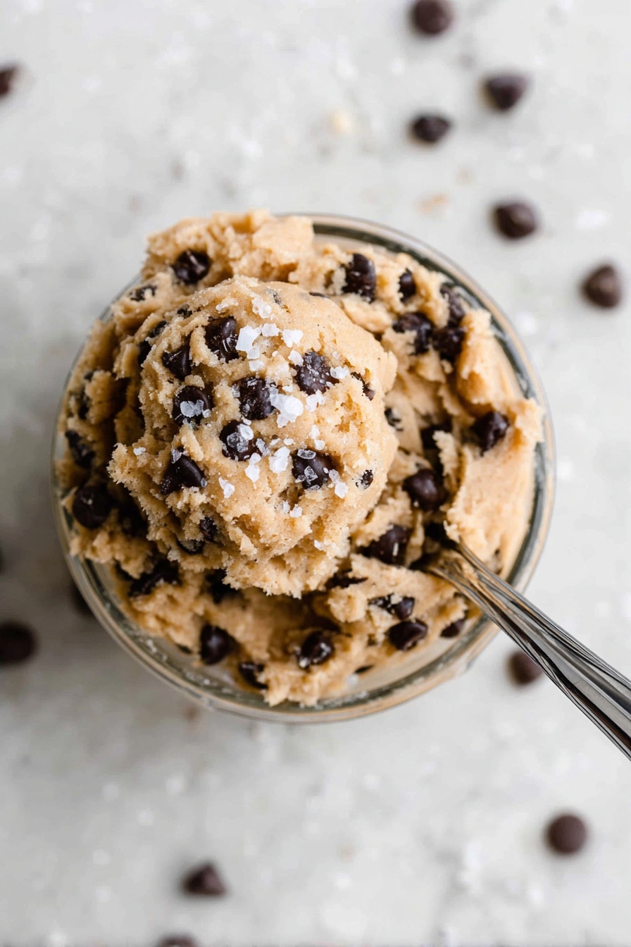 Protein Cookie Dough Bites, healthy protein snacks, no-bake protein treats, high-protein dessert ideas, easy healthy snack recipes - In a small clear glass bowl filled with one layer of light brown, soft cookie dough mixed with many small dark chocolate chips all over, the dough is slightly scooped at the top with a few coarse white salt flakes sprinkled on it. A shiny silver spoon is stuck into the dough from the right side, angled toward the top right corner, resting inside the bowl. The bowl sits on a surface with a white marbled texture, and some chocolate chips are scattered around it. The photo is taken with an iphone --ar 2:3 --v 7