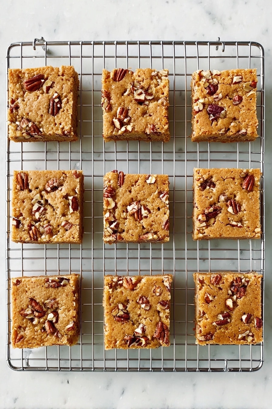 Maple Pecan Blondies, Maple Pecan Blondies recipe, how to make blondies, easy blondie desserts, nutty maple blondies - Nine square pieces of light brown nut bars sit on a silver cooling rack. Each bar has small, uneven chunks of pecans scattered on top, creating a rough texture. The bars are evenly spaced in three rows and three columns, showing a soft, baked surface. The background is a white marbled texture. Photo taken with an iphone --ar 2:3 --v 7