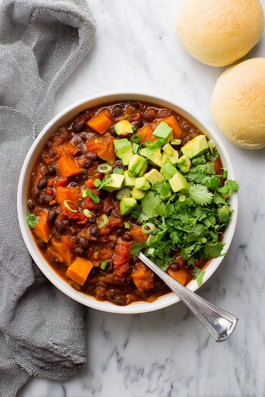 Sweet Potato Black Bean Chili, hearty vegan chili, healthy chili recipe, easy vegetarian chili, comforting plant-based dinner - A white bowl filled with three main layers: the base is a thick stew of dark brown beans mixed with deep orange chunks of sweet potato and red tomato pieces in a reddish-brown sauce, topped on one side with bright green chopped avocado cubes and sprinkled with small rings of green onion, and on the other side with fresh green cilantro leaves. A silver spoon rests inside the bowl at the right side. The bowl sits on a white marbled surface next to two light beige round bread rolls and a loosely folded gray cloth. Photo taken with an iphone --ar 2:3 --v 7