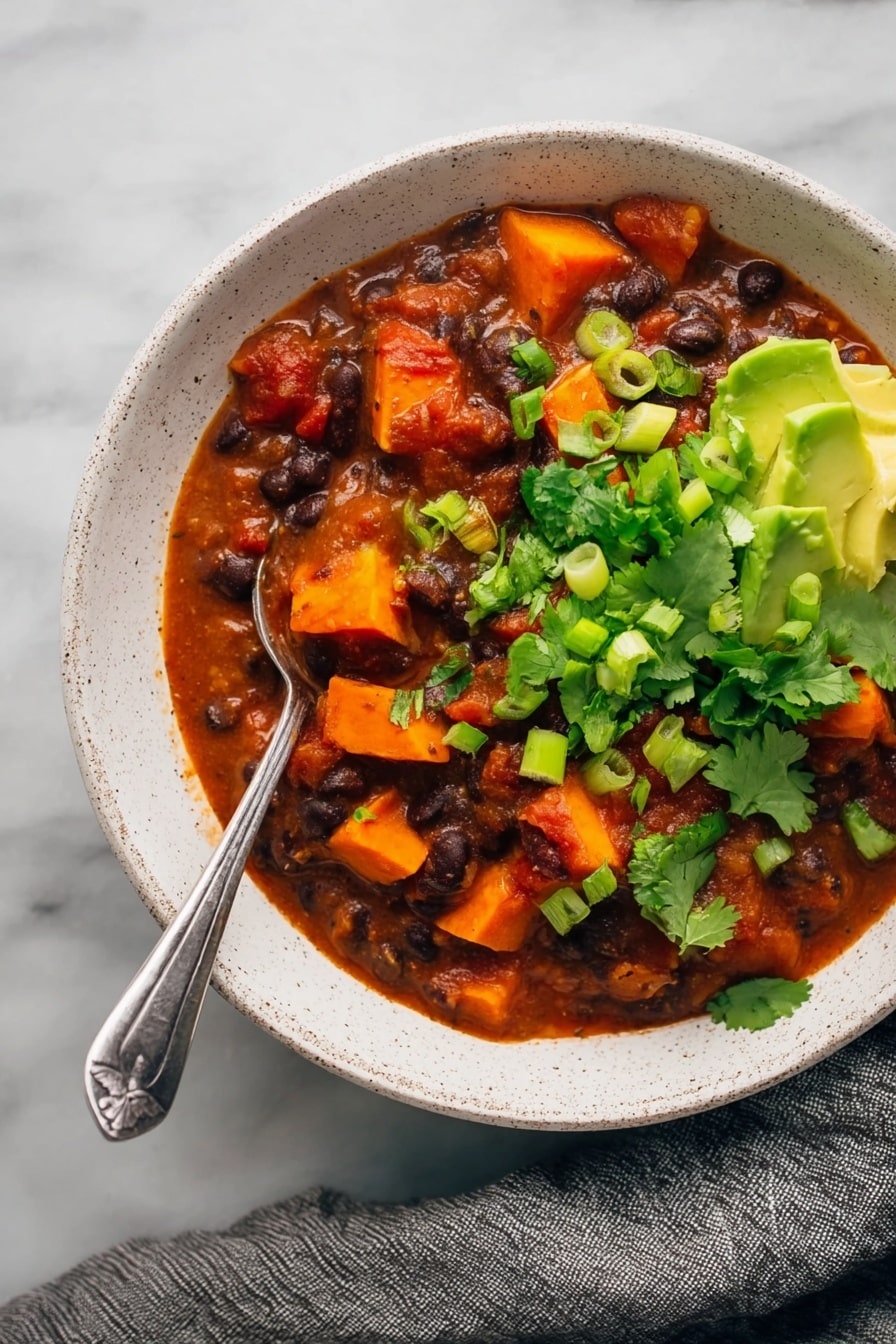 Sweet Potato Black Bean Chili, hearty vegan chili, healthy chili recipe, easy vegetarian chili, comforting plant-based dinner - The image shows a white speckled bowl filled with a thick stew made of dark black beans and bright orange chunks of sweet potato in a rich red tomato sauce. The stew is garnished with vibrant fresh green cilantro leaves and sliced green onions, while creamy green avocado pieces sit on one side near the bowl’s edge. A silver spoon is placed in the stew with its handle resting on the edge of the bowl. The bowl is set on a white marbled surface with a gray cloth draped nearby. photo taken with an iphone --ar 2:3 --v 7