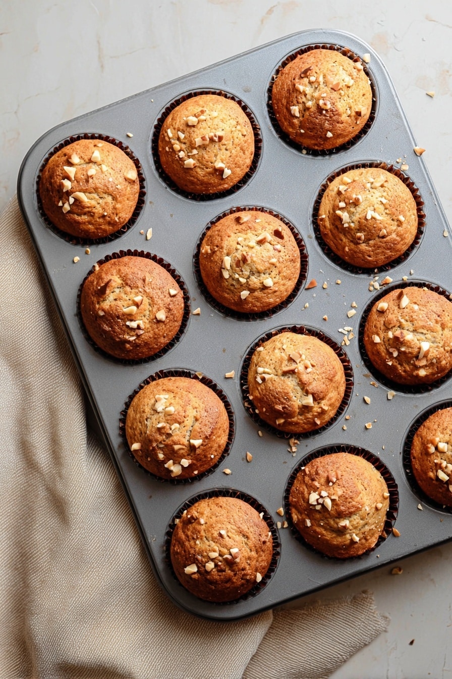 Quick & Easy Vegan Banana Muffins, vegan banana muffins, healthy banana muffin recipe, gluten-free banana muffins, breakfast banana muffins - A grey muffin tray holds twelve freshly baked muffins, each with a golden-brown top that is slightly rounded and textured with small cracks. Light tan chopped nuts are scattered on the surface of each muffin, adding a crunchy detail. The muffins sit in dark brown paper liners that are barely visible around the edges. The tray rests on a white marbled texture surface with a soft beige cloth partially visible on the left side. The light gently highlights the warm tones and textures of the muffins, making them look soft and fresh. photo taken with an iphone --ar 2:3 --v 7