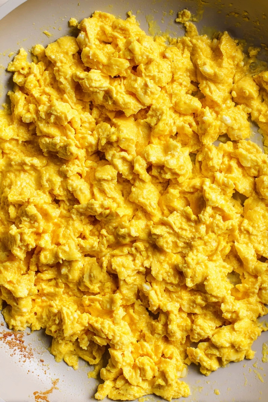 The Best Tofu Scramble, vegan breakfast recipes, healthy tofu scramble, easy plant-based breakfast, protein-packed tofu scramble - The image shows a close-up view inside a white pan filled with scrambled eggs. The cooked eggs have a soft, slightly chunky texture with a bright yellow color spread evenly across the pan. The edge of the pan reveals some lightly browned bits where the eggs touched the surface. The background is a white marbled texture. photo taken with an iphone --ar 2:3 --v 7