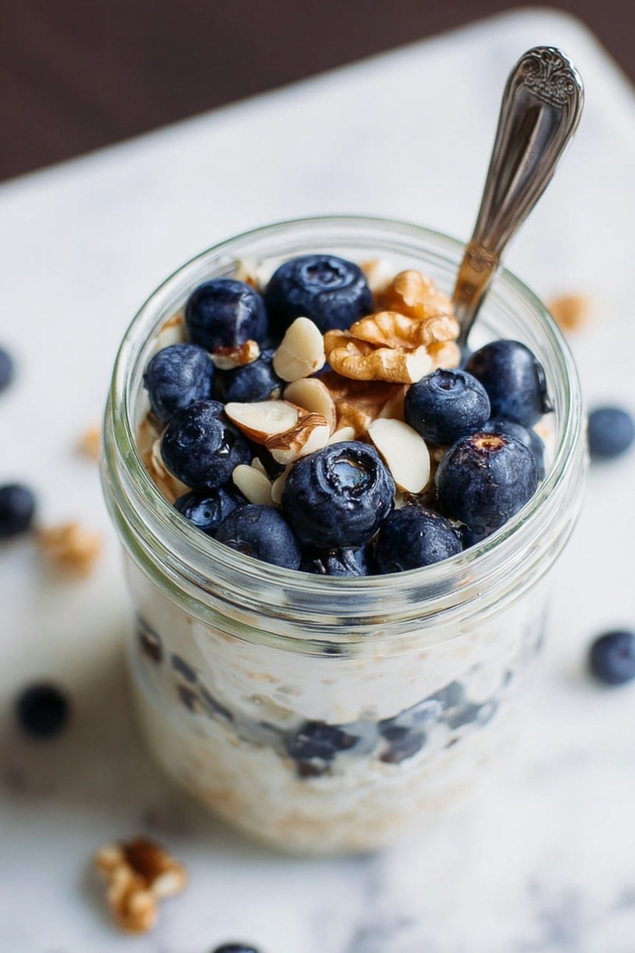 Simple Vegan Overnight Oats, vegan breakfast ideas, healthy overnight oats, easy plant-based breakfast, customizable vegan oats - A clear glass jar filled with three layers: the bottom layer is fresh blueberries, the middle layer is creamy white porridge or yogurt with a smooth texture, and the top layer has more blueberries and a few light brown walnut pieces scattered on top. A silver spoon is placed in the jar, partly inside the food. The surface around the jar shows a white marbled texture with some scattered white nut pieces. Photo taken with an iphone --ar 2:3 --v 7