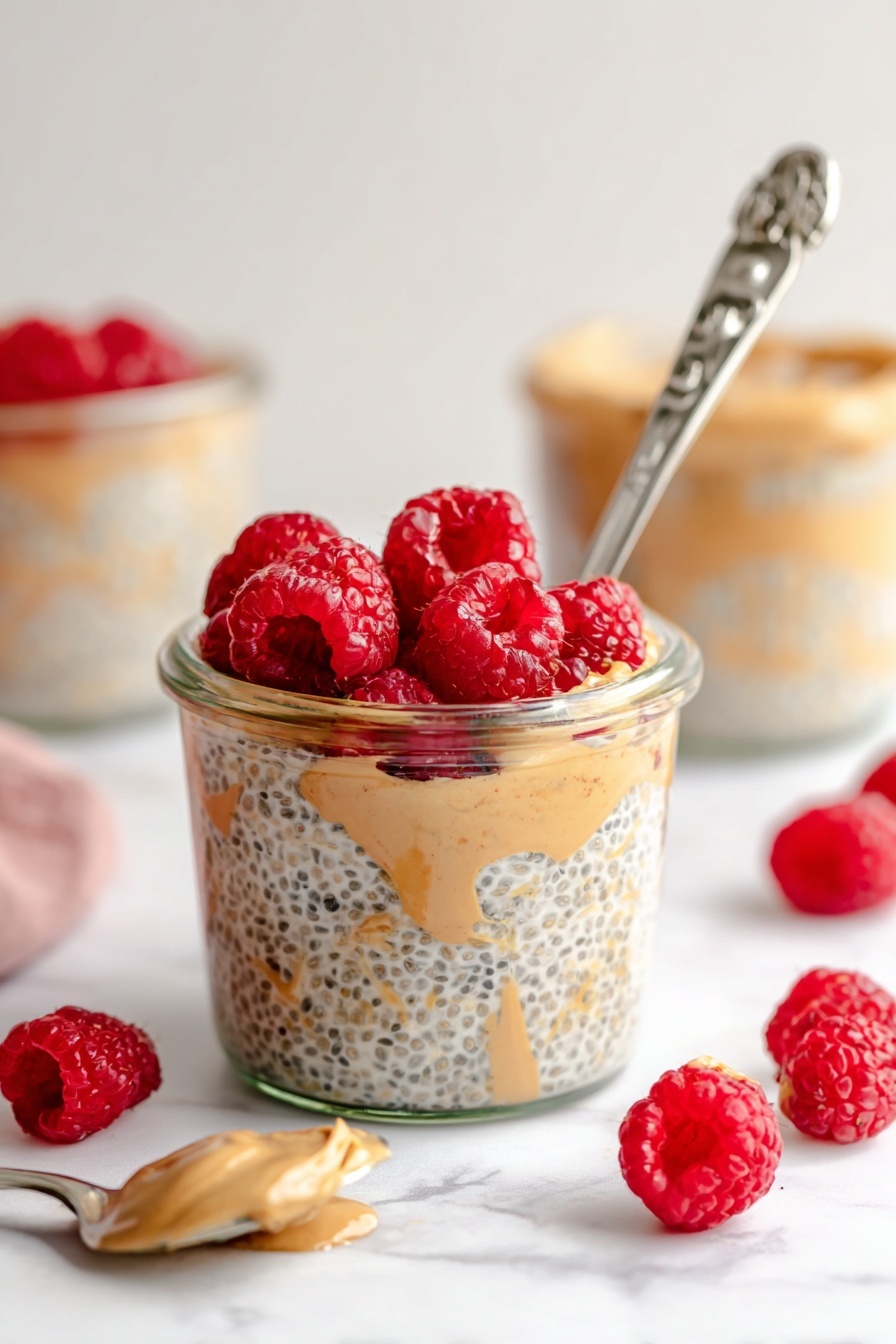 Peanut Butter Overnight Oats, healthy overnight oats, easy breakfast recipes, quick morning breakfast, nutritious oatmeal - A clear glass jar filled with three visible layers: the bottom layer is light beige chia seed pudding with black chia seeds mixed in, the middle layer shows swirls of creamy light brown peanut butter, and the top layer is a bright red cluster of fresh raspberries. A silver spoon with an ornate handle is placed inside the jar, leaning against the side with some pudding on it. Around the jar, scattered fresh raspberries add more red spots on a white marbled surface. In the background, more jars with similar contents and a silver spoon with a light brown spread are softly blurred. Photo taken with an iphone --ar 2:3 --v 7