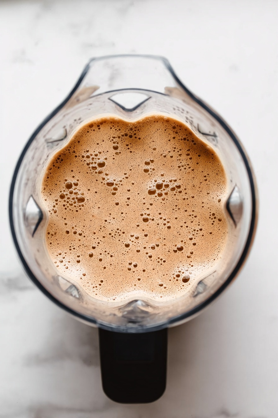 Vegan Chocolate Banana Protein Shake, healthy vegan smoothie, quick plant-based protein shake, chocolate banana smoothie, nutritious vegan breakfast - The image shows a clear blender from above filled with a light brown, frothy mixture with many small bubbles on its surface. The blender has a black handle, and the background is a white marbled surface. photo taken with an iphone --ar 2:3 --v 7
