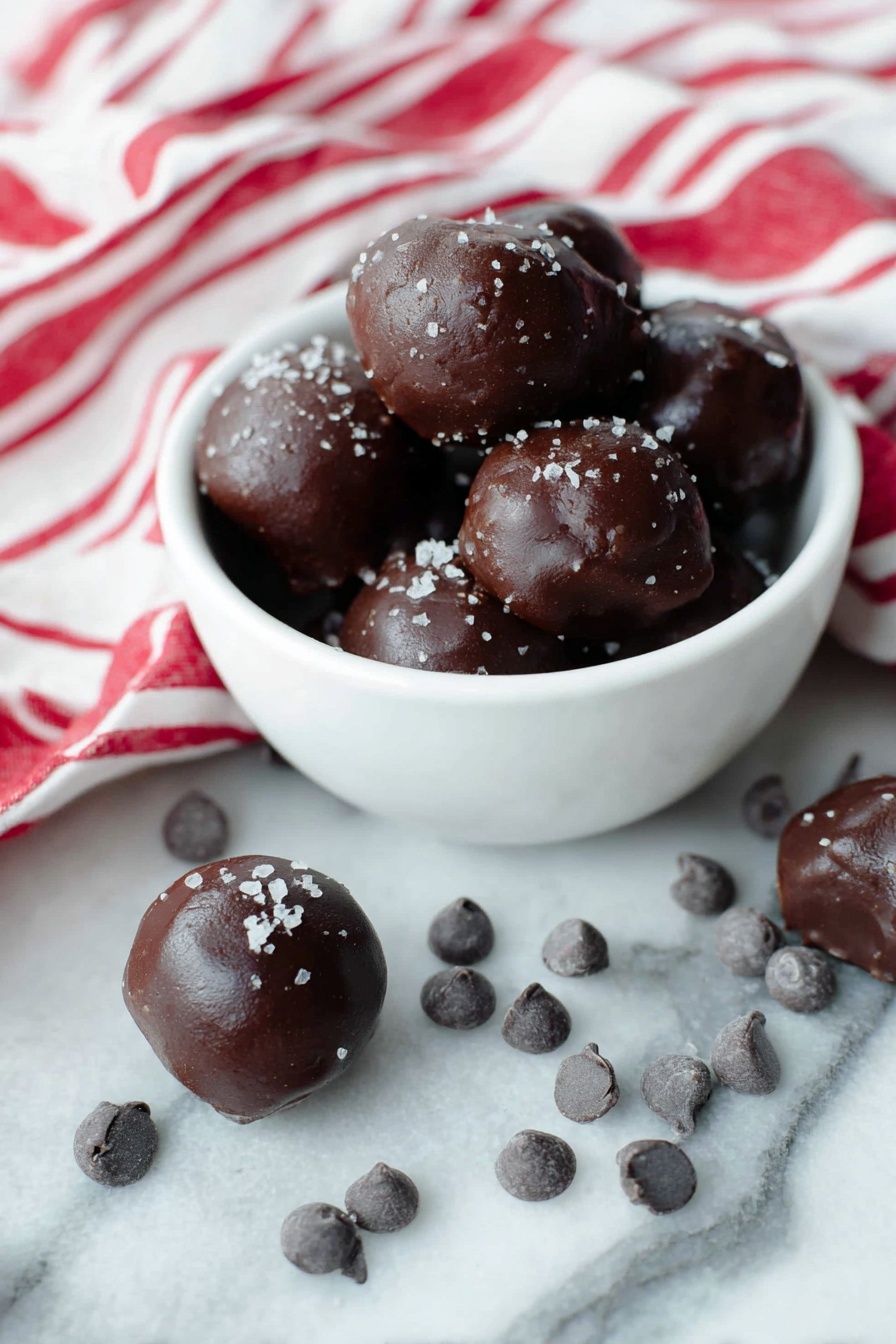 Cocoa Date Truffles, healthy date truffles, no-bake chocolate snacks, vegan energy bites, simple dessert ideas - A white bowl filled with several dark brown chocolate balls that have a smooth, shiny texture and are sprinkled lightly with coarse salt flakes on top. Around the bowl, more chocolate balls sit on a white marbled surface, some resting near small clusters of dark chocolate chips. A white cloth with bold red stripes is casually draped to the back and right side of the bowl, adding a bright contrast. The scene is bright and clean, showing the rich dark color and glossy texture of the chocolates clearly. Photo taken with an iphone --ar 2:3 --v 7