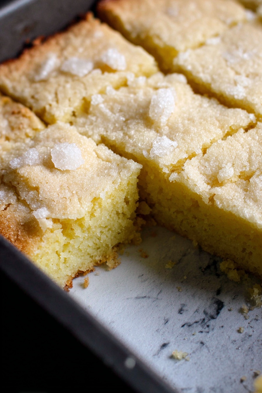 Fluffy French Butter Cake, French Butter Cake Recipe, Easy French Cake, Tender Butter Cake, French Dessert - A single-layer light yellow cake is shown inside a dark baking pan with a white marbled texture surface underneath. The top layer has a rough crumbly texture with sugar crystals scattered on top, and it is cut into square pieces with one piece missing, revealing a soft, dense inside that is smooth and slightly moist. The cake edges are slightly browned and cracked, contrasting with the pale yellow center. Photo taken with an iphone --ar 2:3 --v 7