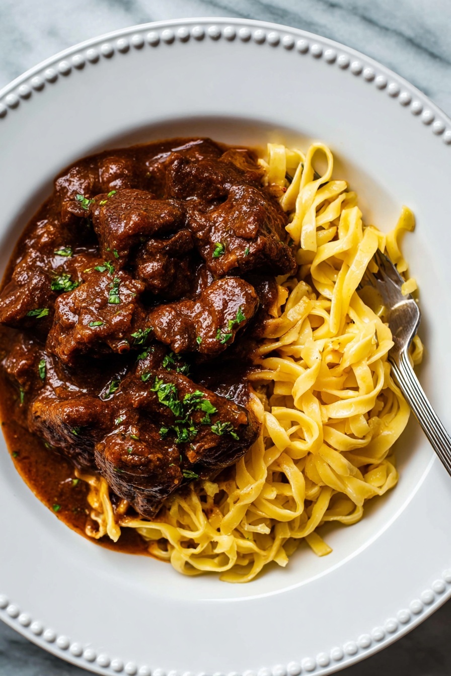 German Beef Goulash, hearty beef goulash recipe, traditional German goulash, slow cooker beef goulash, easy beef goulash dinner - A white plate with a raised dotted edge holds a dish of thick yellow noodles on the bottom right, with a silver fork resting on them. On top and mostly to the left of the noodles, there are large dark brown chunks of meat in rich brown sauce. The sauce is thick and covers most of the meat, with small pieces of green herbs sprinkled over the meat and noodles. The plate is placed on a white marbled surface. photo taken with an iphone --ar 2:3 --v 7