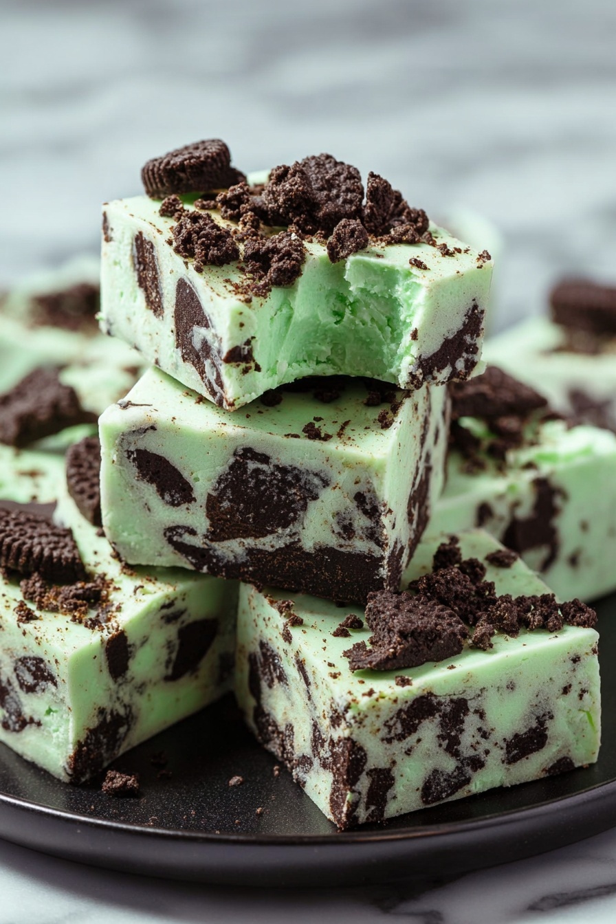 Mint Oreo White Chocolate Fudge, mint Oreo fudge, white chocolate fudge recipes, no-bake fudge desserts, creamy mint fudge - The image shows a stack of five square pieces of mint chocolate fudge on a black plate, placed on a white marbled surface. The fudge has a pale green base with dark brown chunks of cookie mixed in all layers. On the top, bigger cookie pieces and crumbs are scattered, adding texture and color contrast. Each piece is thick and smooth with distinct edges, and the top piece has a small bite taken from its corner, showing a creamy interior with visible cookie pieces inside. Photo taken with an iphone --ar 2:3 --v 7