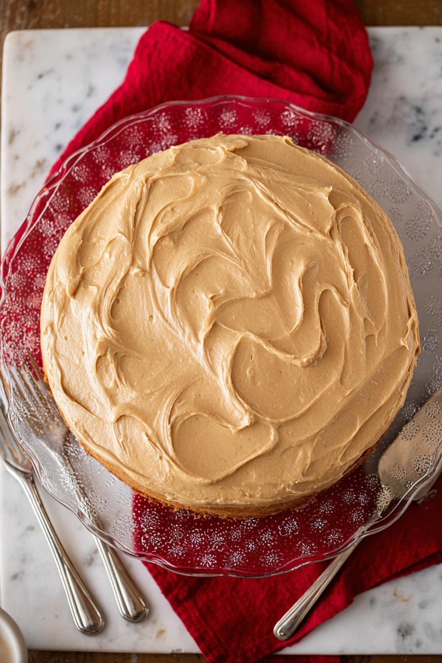 Apple Spice Cake with Caramel Icing, cozy apple cake recipe, autumn apple spice cake, homemade caramel icing, moist apple cake dessert - A round cake with one visible layer is covered evenly with smooth light brown frosting that has soft swirls on top. The cake sits on a clear glass plate with a floral pattern, placed on a white marbled surface with a red cloth underneath the plate. Two silver forks are on the left side of the plate, and a silver cake server is on the right side. The scene is well-lit, showing the texture of the frosting clearly photo taken with an iphone --ar 2:3 --v 7