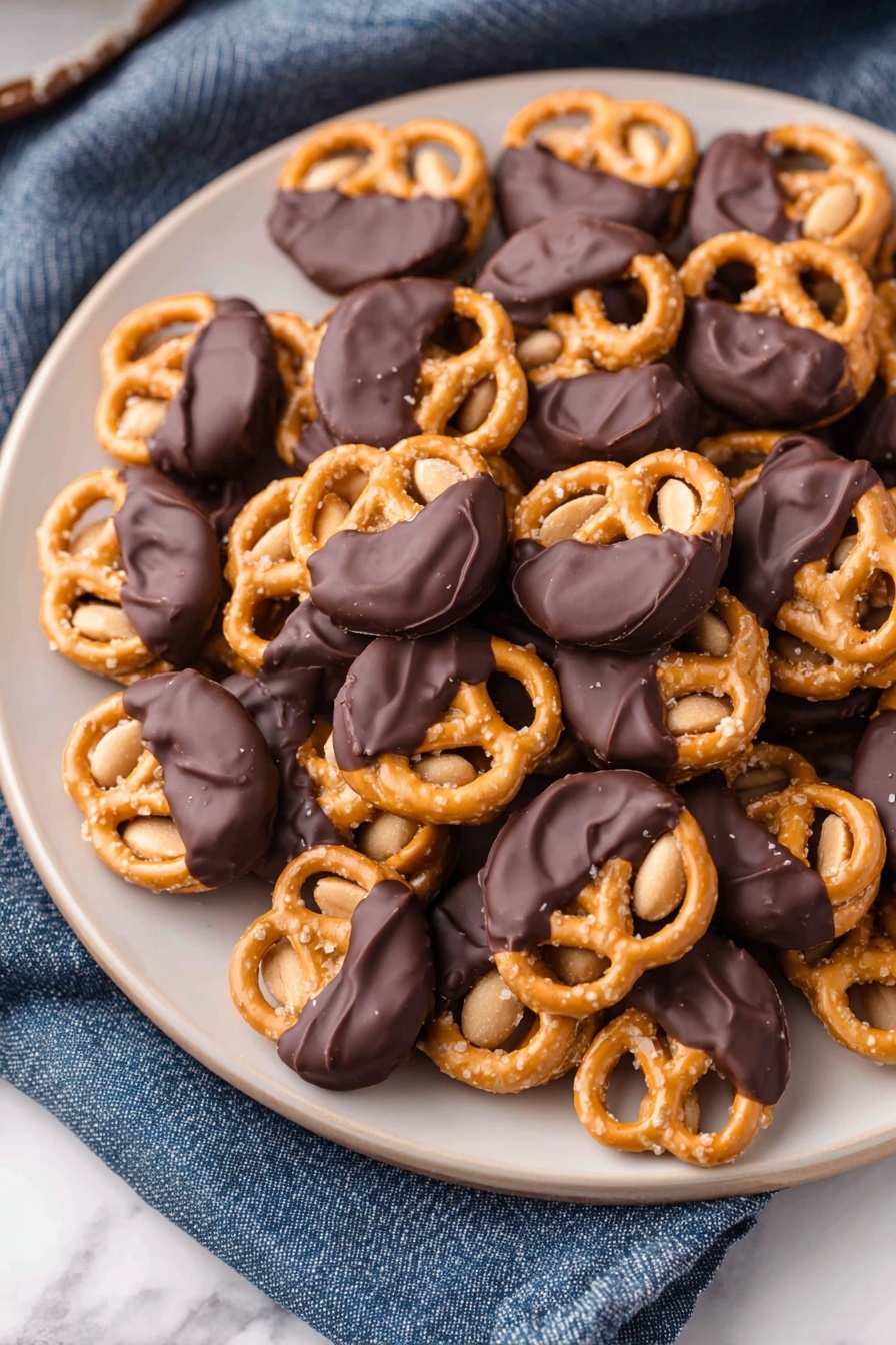 Chocolate Dipped Peanut Butter Pretzel Bites, peanut butter pretzel treats, salty sweet snack recipes, easy dessert bites, chocolate dipped pretzels - A white plate filled with many small pretzels, each topped with a single peanut and half dipped in smooth dark chocolate, giving a mix of shiny black and golden tan colors. The pretzels have a crunchy texture with visible salt grains on the surface, while the peanuts underneath add a light brown soft contrast. The plate sits on a white marbled texture with a blue cloth nearby. photo taken with an iphone --ar 2:3 --v 7