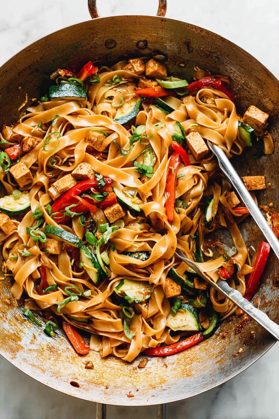 Drunken Noodles with Chicken, Thai Drunken Noodles Recipe, Spicy Chicken Noodles Thai, Easy Thai Chicken Noodles, Authentic Thai Noodles Recipe - The image shows a metal wok filled with a colorful stir-fry dish. The base layer consists of wide, flat noodles that are light brown and glossy. Mixed through the noodles are pieces of lightly browned tofu cubes, thin strips of green bell peppers, slices of red bell peppers, and small chunks of zucchini with their green skin visible. Scattered over the top are thinly sliced green onions and bits of chopped herbs adding dots of bright green. The noodles and vegetables appear shiny from a light sauce coating them evenly, and the wok has a warm, well-used appearance with some browning on the sides. A pair of metal tongs rests inside the wok, ready to serve. The setting has a clean, white marbled texture surface. photo taken with an iphone --ar 2:3 --v 7