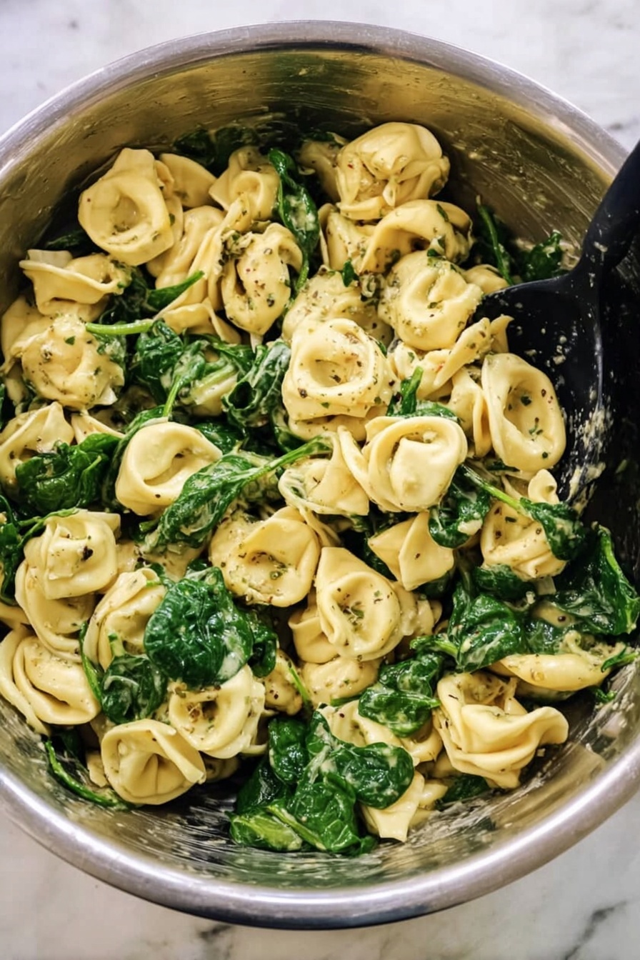 Creamy Pesto Tortellini with Spinach, easy pesto tortellini, quick weeknight pasta dinner, vegetarian pasta recipes, flavorful cheese tortellini - This image shows a big metal bowl filled with cooked tortellini pasta mixed with bright green spinach leaves. The pasta is light yellow with a soft texture, and some pieces have dark green herb specks on them. The spinach looks fresh and slightly wilted, spread evenly throughout the pasta. There is a black spoon resting inside the bowl, ready to serve. The bowl sits on a white marbled surface, giving a clean and simple background. Photo taken with an iphone --ar 2:3 --v 7