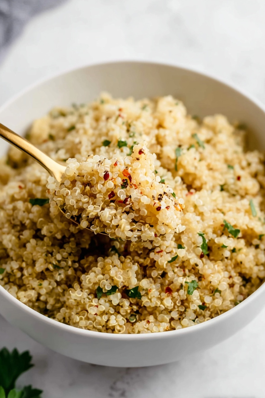 Garlic Butter Quinoa, quick healthy side dish, easy quinoa recipes, flavorful side meals, garlic butter recipe - A close-up of a white bowl full of cooked quinoa, which is light beige and fluffy. The quinoa grains are mixed with small green herb pieces scattered evenly throughout and seasoning specks of black and red on top. A gold spoon scoops up some quinoa from the bowl, showing the grain’s soft, slightly textured surface. The bowl sits on a white marbled texture surface with some green leaves blurred in the background. photo taken with an iphone --ar 2:3 --v 7