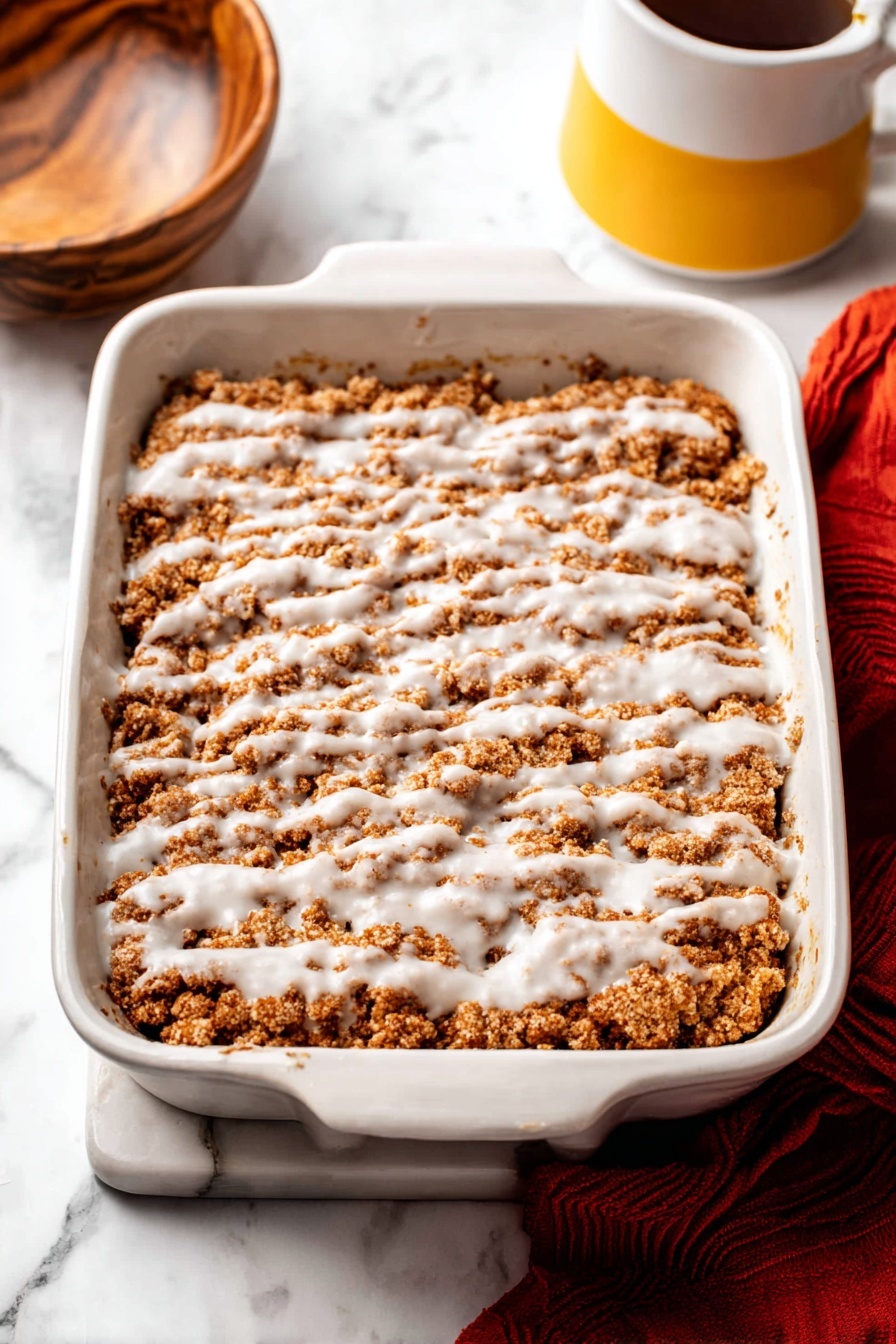 Pumpkin Crumb Coffee Cake, pumpkin coffee cake with crumb topping, fall coffee cake recipe, easy pumpkin coffee cake, moist pumpkin coffee cake - The image shows a white rectangular baking dish filled with a crumbly brown dessert layer, topped with a drizzle of white icing spread unevenly across the surface. The crumb layer has a soft, rough texture with patches of darker and lighter brown. The dish is placed on a white marbled surface, with part of a wooden bowl visible on the left and a yellow and white mug on the right. A red cloth lies partially beneath the baking dish at the bottom right corner. Photo taken with an iphone --ar 2:3 --v 7