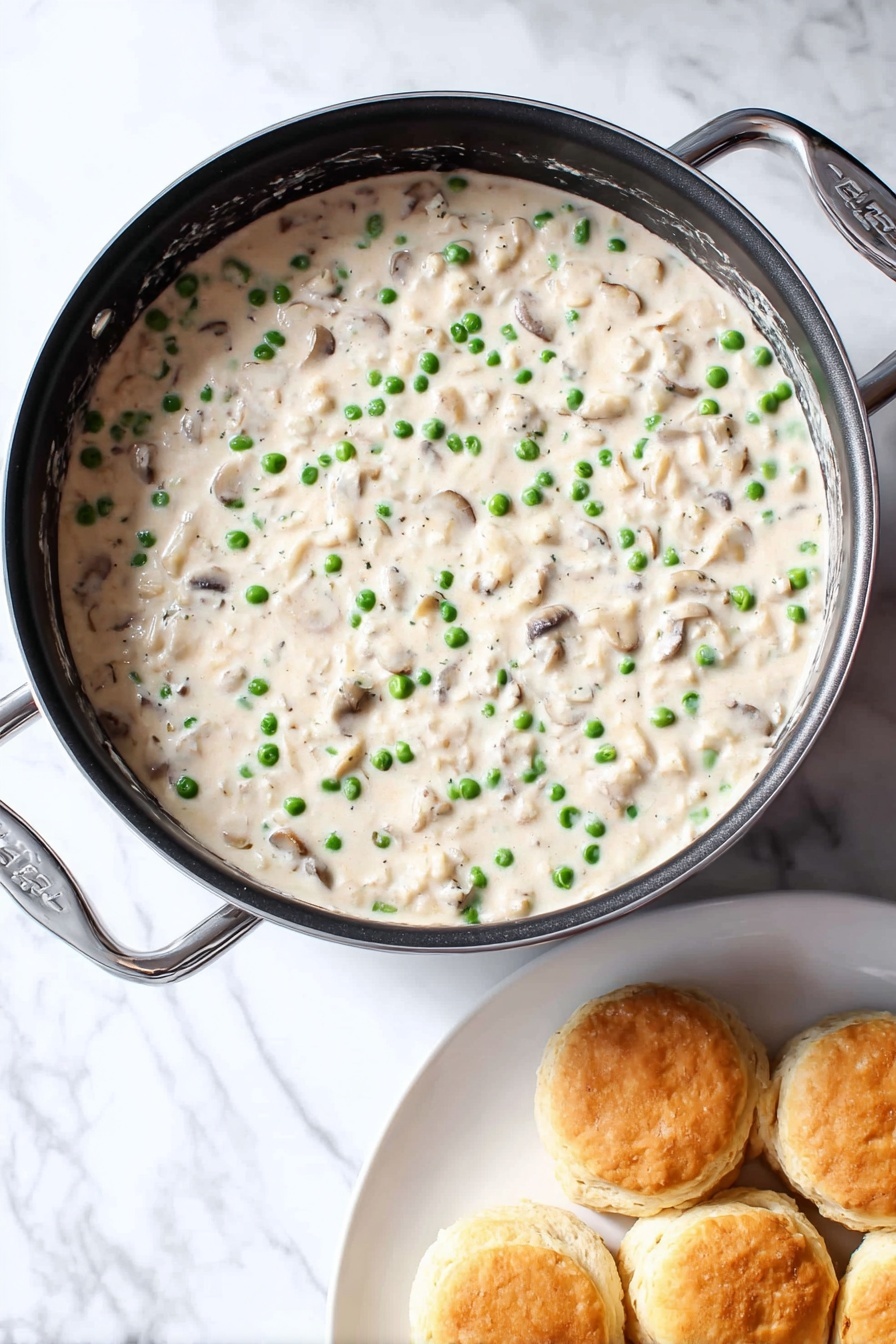 Creamed Chicken Over Biscuits, Creamed Chicken recipe, savory chicken and biscuits, comforting chicken dinner, easy weekend brunch ideas - A round black pan filled with creamy white sauce that has small green peas and small light brown mushroom pieces mixed evenly throughout. The sauce looks thick and smooth. Next to the pan is a white plate holding about six golden brown biscuits stacked loosely. All is placed on a white marbled surface. photo taken with an iphone --ar 2:3 --v 7