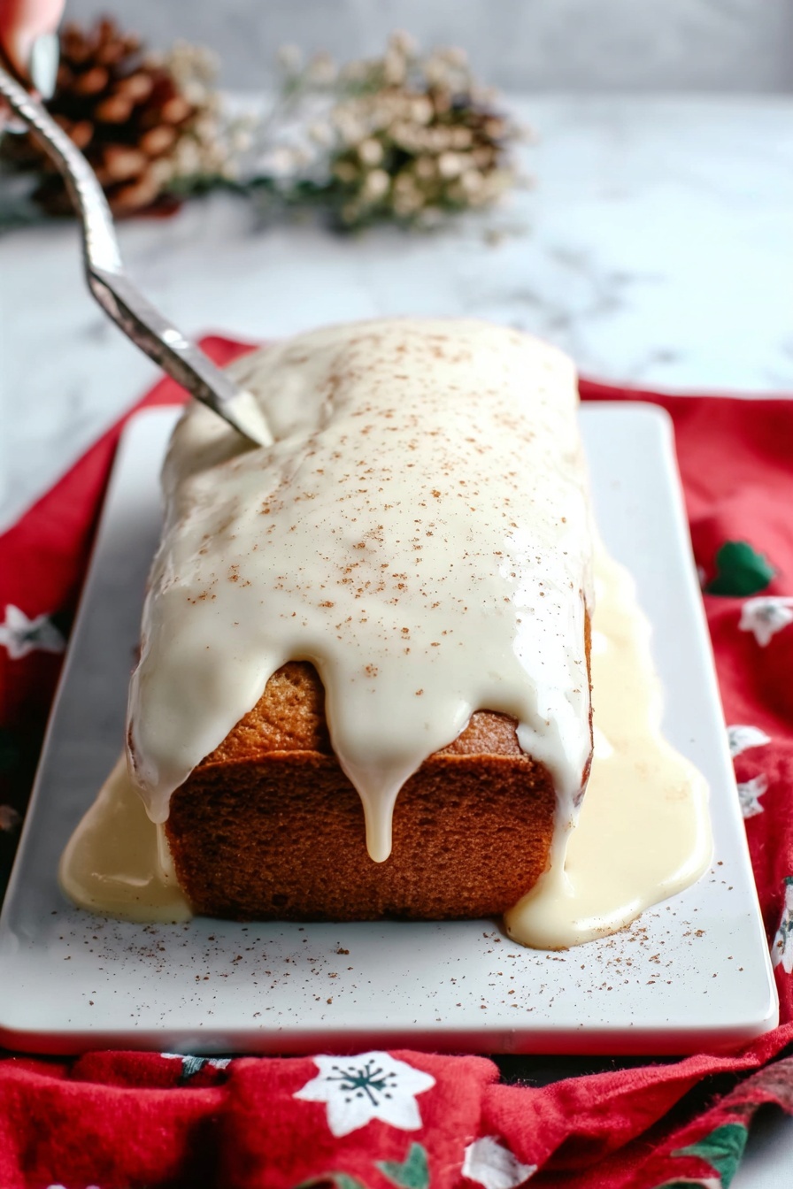 Eggnog Bread Christmas Bread Holiday Sweet Bread Festive Baking - A rectangular loaf cake sits on a white rectangular tray over a red cloth with white and blue flower patterns, placed on a white marbled surface. The cake has a golden-brown color and is covered on top with thick, creamy white icing that drips over the edges and pools around the base. The icing is lightly sprinkled with a fine brown powder, likely cinnamon or nutmeg. The scene includes dried flowers blurred in the background and a woman's hand holding a silver knife positioned above the cake. Photo taken with an iphone --ar 2:3 --v 7