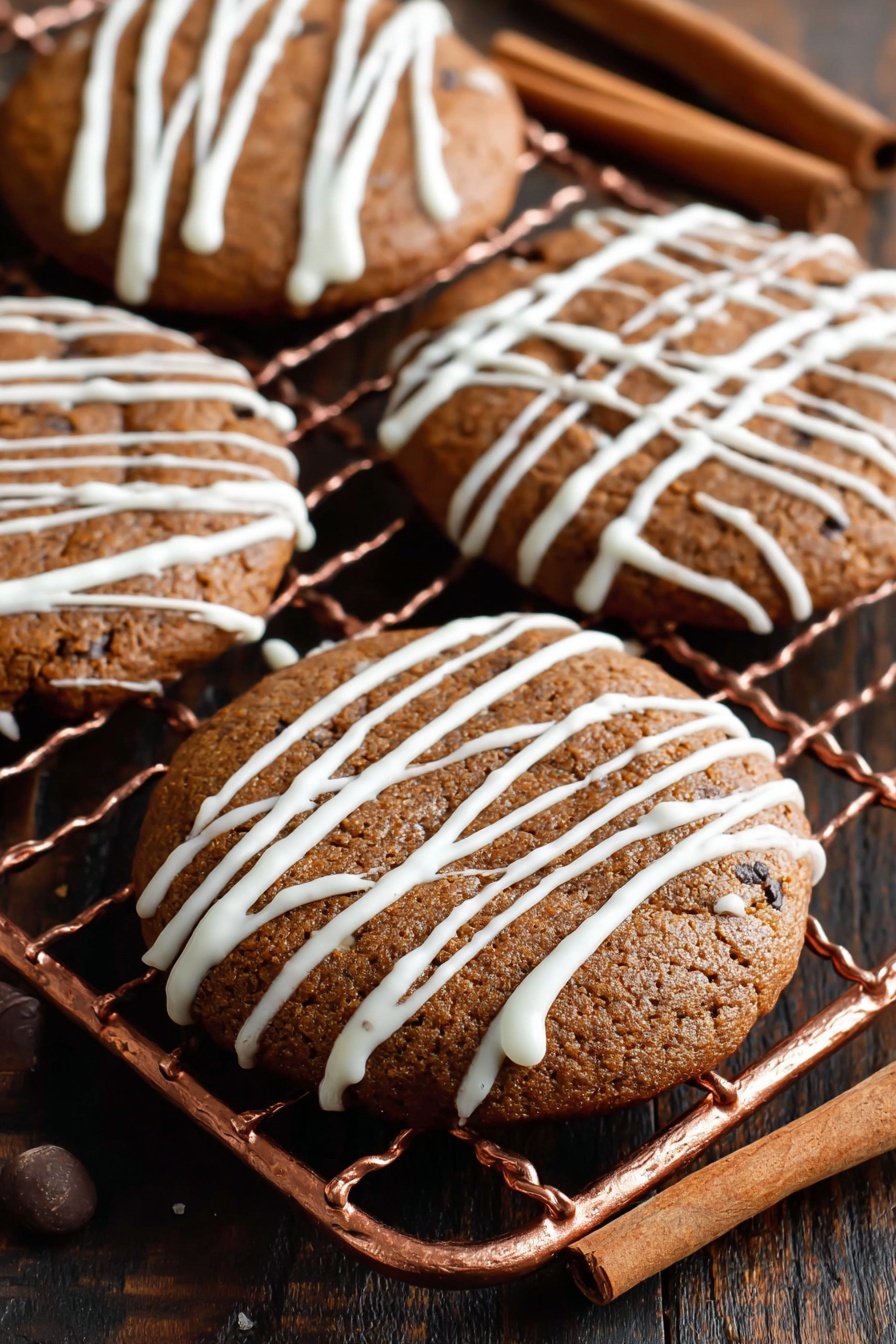 Soft Gingerbread Cookies, Gingerbread Cookies, Festive Gingerbread Cookies, Easy Gingerbread Cookies, Soft Gingerbread Cookies Recipe - The image shows four round brown cookies with a slightly cracked surface, placed on a copper cooling rack. Each cookie is decorated with thin, white icing drizzled randomly across the top, creating a light pattern. The cookies look soft and moist with dark spots indicating spices or chocolate chips. The background includes a dark wooden table with two cinnamon sticks placed on the right side. photo taken with an iphone --ar 2:3 --v 7