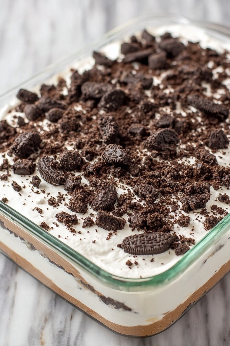 Red Velvet Poke Cake with Cheesecake Pudding, red velvet poke cake, cheesecake pudding dessert, easy red velvet cake, no-bake cheesecake poke cake - A clear glass rectangular dish holds a layered dessert with three visible layers. The bottom layer is a light brown creamy base, covered by a thick middle layer of smooth white cream. The top layer is sprinkled generously with roughly crushed dark chocolate sandwich cookies, scattered evenly across the white cream. The dish sits on a white marbled surface. photo taken with an iphone --ar 2:3 --v 7