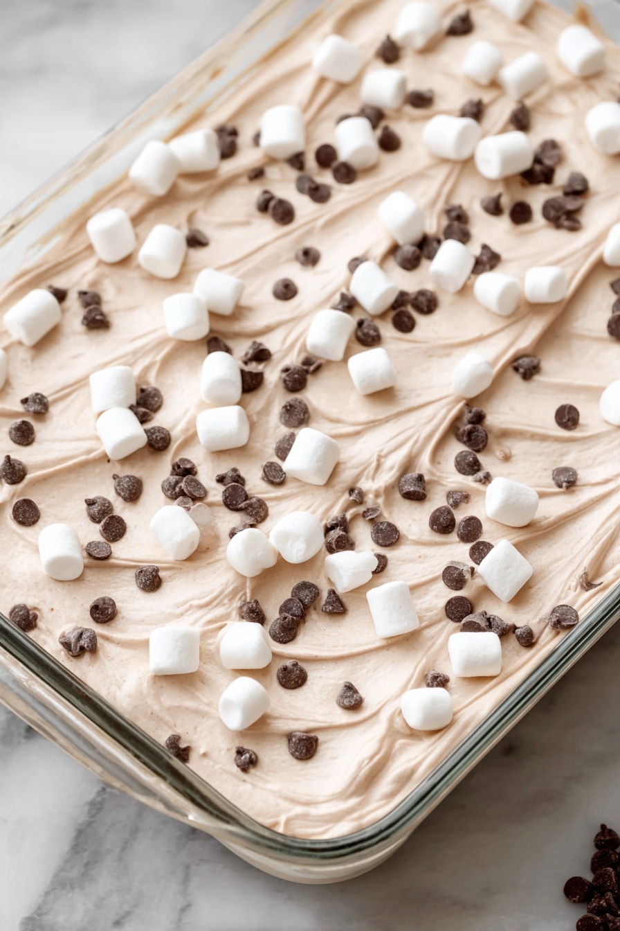 Hot Chocolate Poke Cake, chocolate poke cake, marshmallow fudge cake, hot chocolate dessert, easy chocolate cake recipe - A dessert in a clear rectangular glass dish with one thick layer of light brown creamy mousse or whipped topping, spread evenly with soft swirls on the surface. Scattered on top are many small white marshmallows along with a good amount of very tiny dark brown chocolate chips spread all over. The dish is placed on a white marbled surface. Photo taken with an iphone --ar 2:3 --v 7