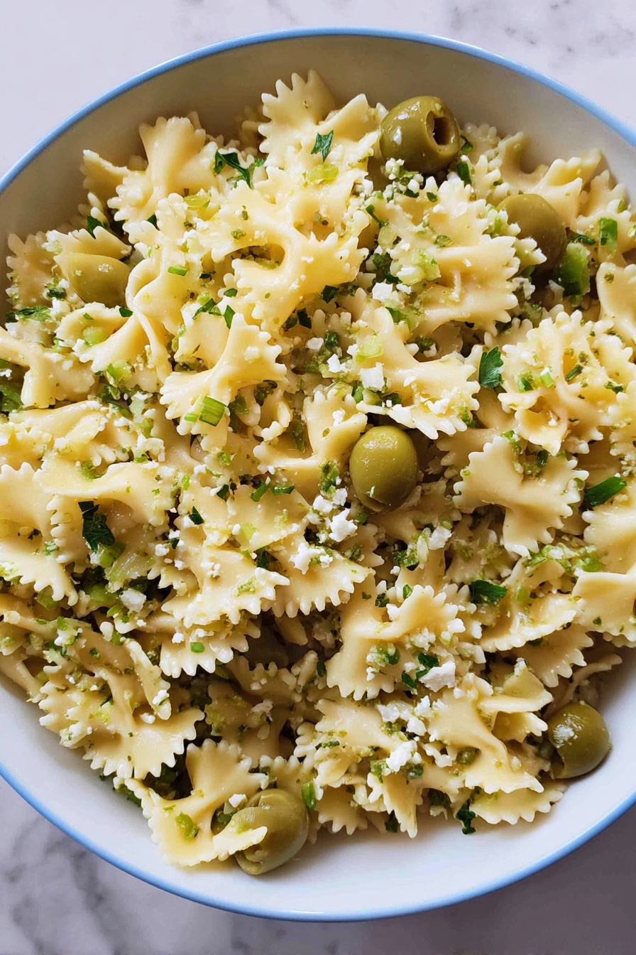 Dirty Martini Pasta Salad, pasta salad with olives and lemon, savory pasta salad recipe, summer pasta salad ideas, bold flavored pasta salad - A close-up view of a large white bowl filled with curly-edged pasta that is light yellow in color, mixed evenly with bright green sliced olives and sprinkled with small bits of light green herbs and crumbled white cheese. The pasta is separated nicely, showing the wavy edges and soft texture, while the green olives add contrast throughout. The bowl sits on a white marbled surface, creating a clean and fresh look. photo taken with an iphone --ar 2:3 --v 7