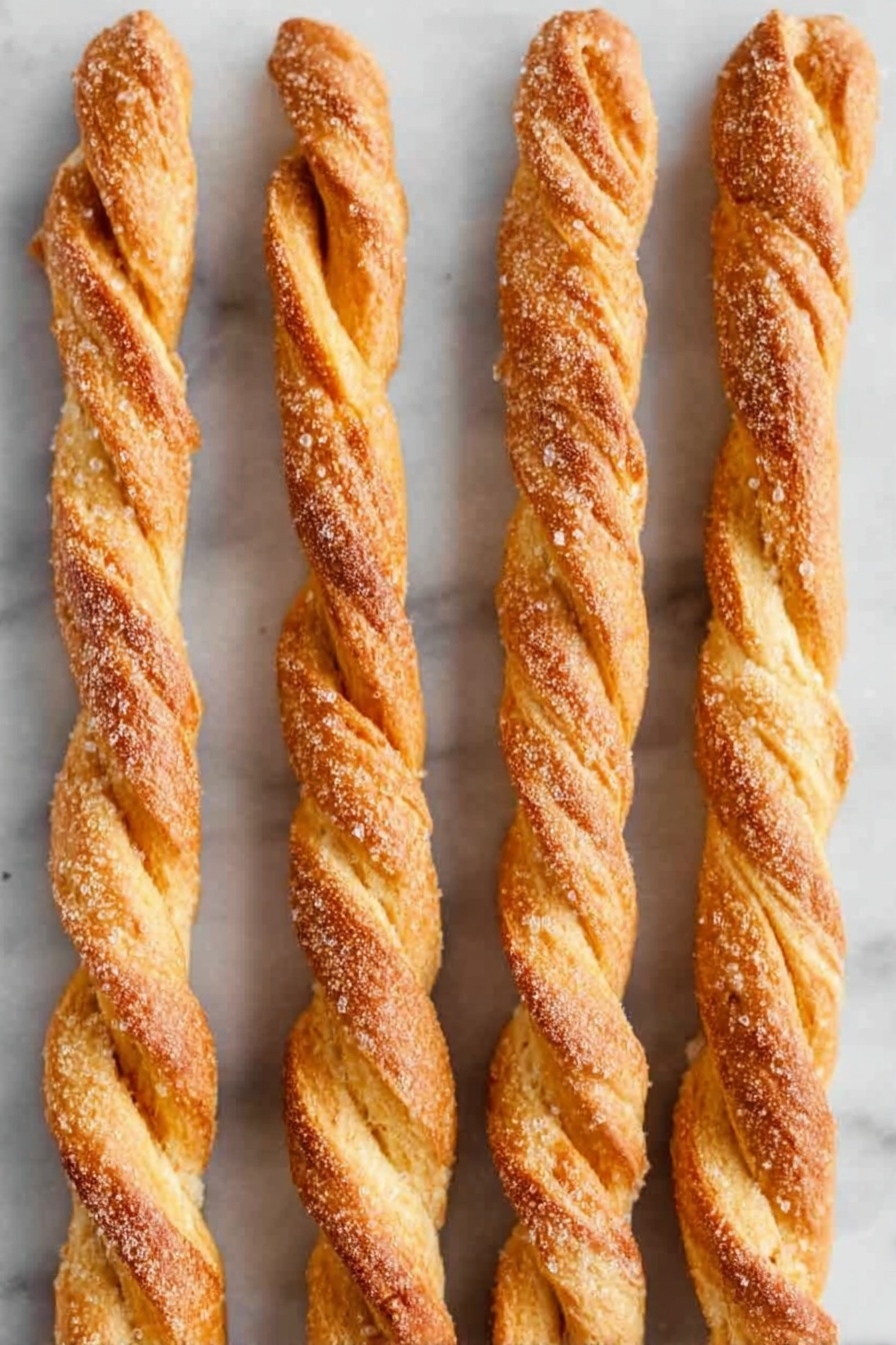 Pumpkin Pie Twists with Crescent Dough, Pumpkin Pie Twists, easy pumpkin pie snacks, autumn dessert ideas, holiday appetizer recipes - The image shows several twisted pastry sticks arranged closely on a white rectangular plate. Each pastry stick has a golden-brown crust with a dusting of cinnamon sugar on top, giving a slightly rough texture from the sugar granules. The twists reveal light, flaky layers with a cinnamon brown filling swirled inside. The background is a white marbled surface, and the pastries look freshly baked with a slight shine from the glaze. photo taken with an iphone --ar 2:3 --v 7