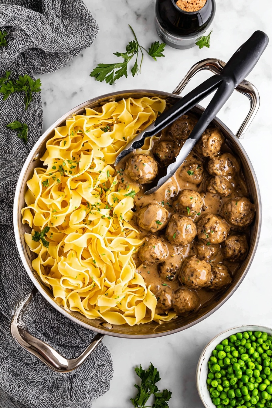 Swedish Meatballs with Creamy Gravy, Swedish meatball recipe, easy Swedish meatballs, homemade Swedish meatballs, comforting meatball dish - A large silver pan with tall handles holds two distinct layers inside: one side is filled with yellow wide pasta noodles twisted and folded over each other with a smooth, slightly shiny texture, while the other side has many round brown meatballs covered in a thick, creamy brown sauce with visible black pepper specks. Metal tongs with black grips rest on top of the pasta, slightly lifting some noodles. The pan is placed on a white marbled surface, surrounded by fresh green parsley leaves scattered around, a gray textured cloth to the left, a white bowl filled with bright green peas on the bottom right, and a dark bottle with cork inside at the top. Photo taken with an iphone --ar 2:3 --v 7