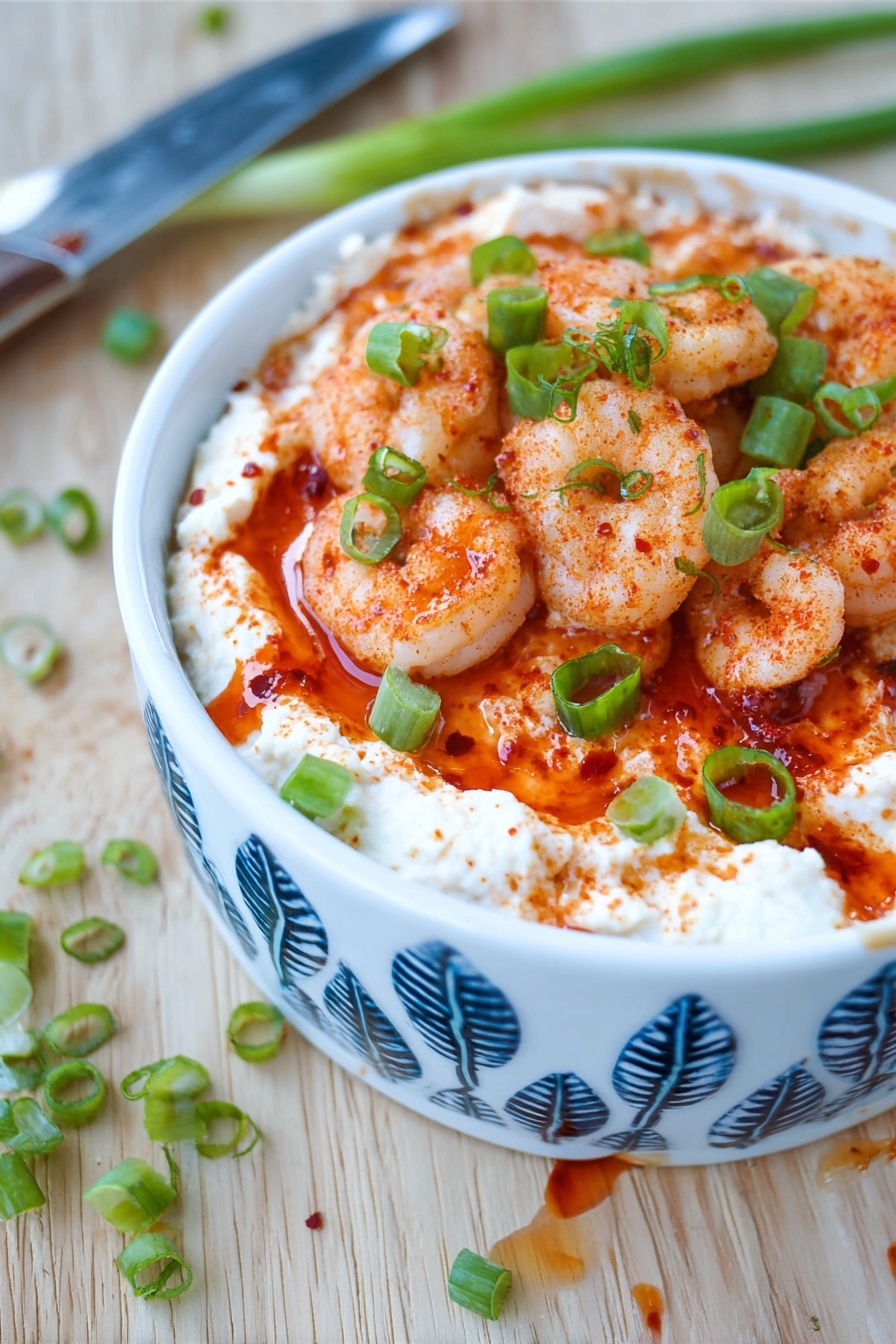 Shrimp Cocktail Dip, Shrimp Dip Recipe, Easy Shrimp Appetizer, Party Dip Ideas, Seafood Dip - The dish shows three layers in a white bowl with blue leaf patterns. The bottom layer is a creamy white spread with a smooth texture. The middle layer is a bright red sauce that looks thick and slightly shiny. The top layer consists of small cooked shrimp, seasoned with red spices giving them a light orange color, and sprinkled with chopped green onions for a fresh green contrast. The bowl is placed on a light wooden surface with a few green onion pieces scattered around and a knife with some sauce on the blade nearby. Photo taken with an iphone --ar 2:3 --v 7